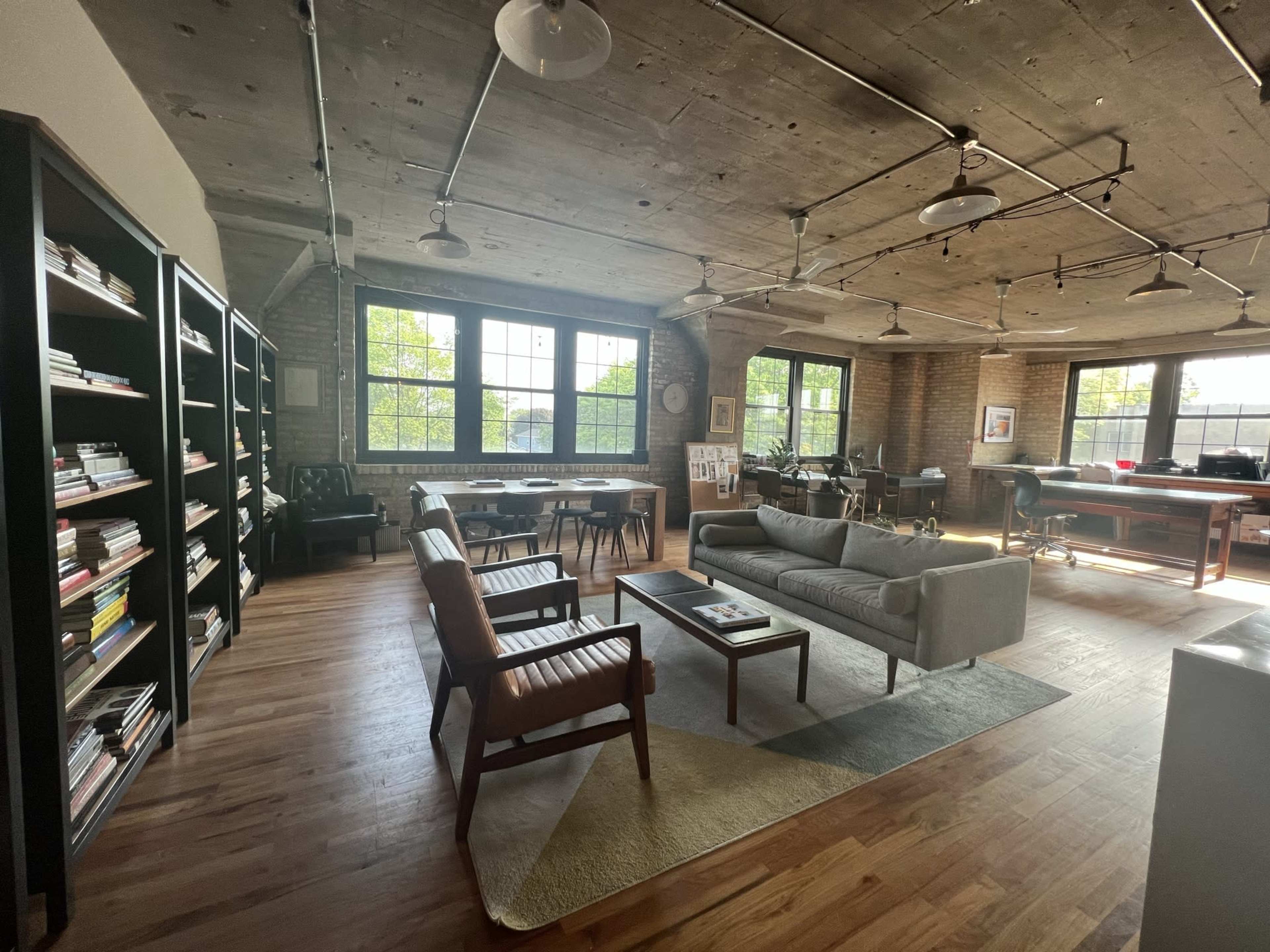 A modern office space features wooden floors, large windows, bookshelves, a seating area with a sofa and chairs, and various workstations.