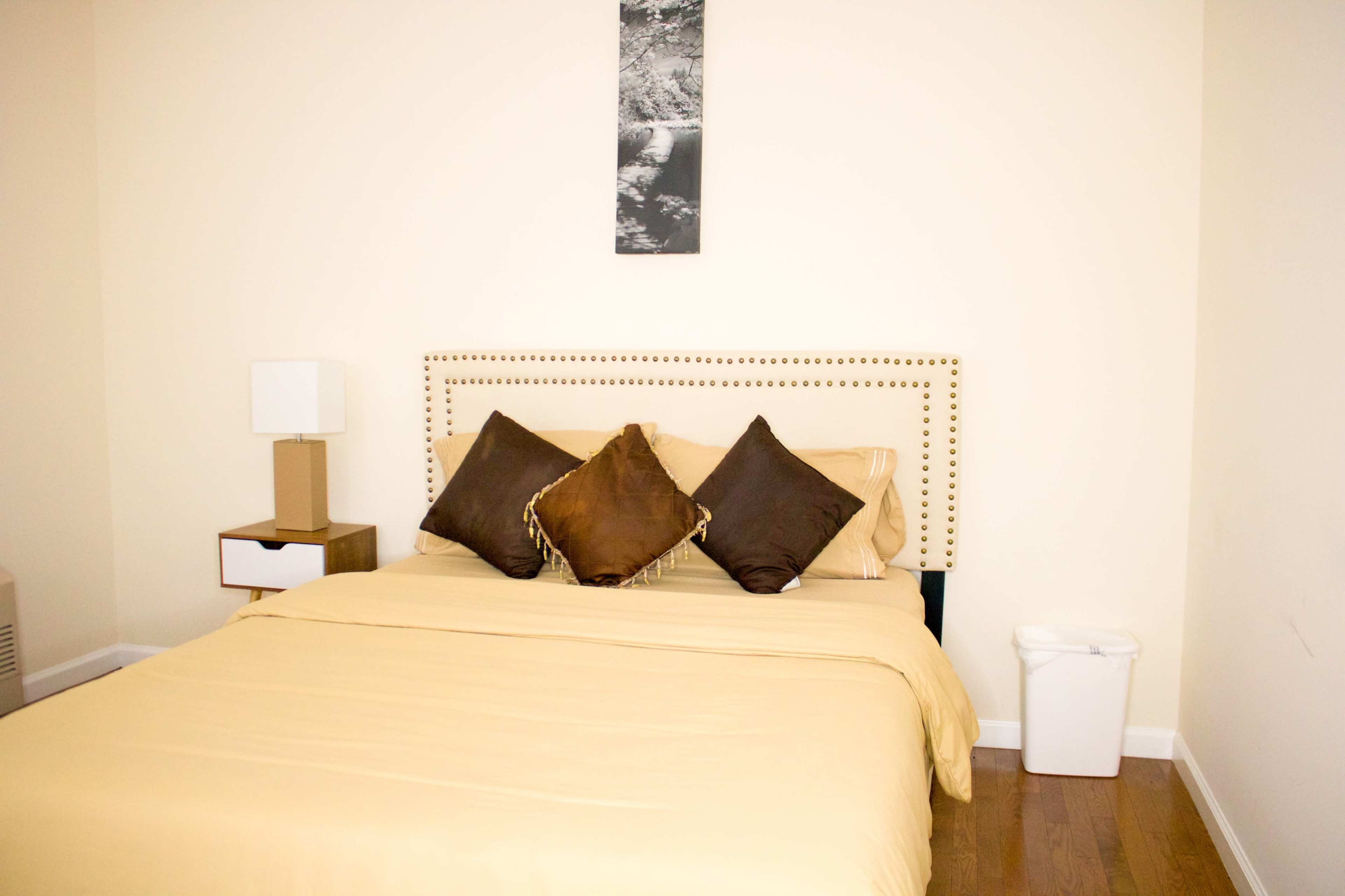 The image shows a neatly made bed with a beige comforter and decorative pillows in a simple bedroom with light-colored walls and minimal furnishings.
