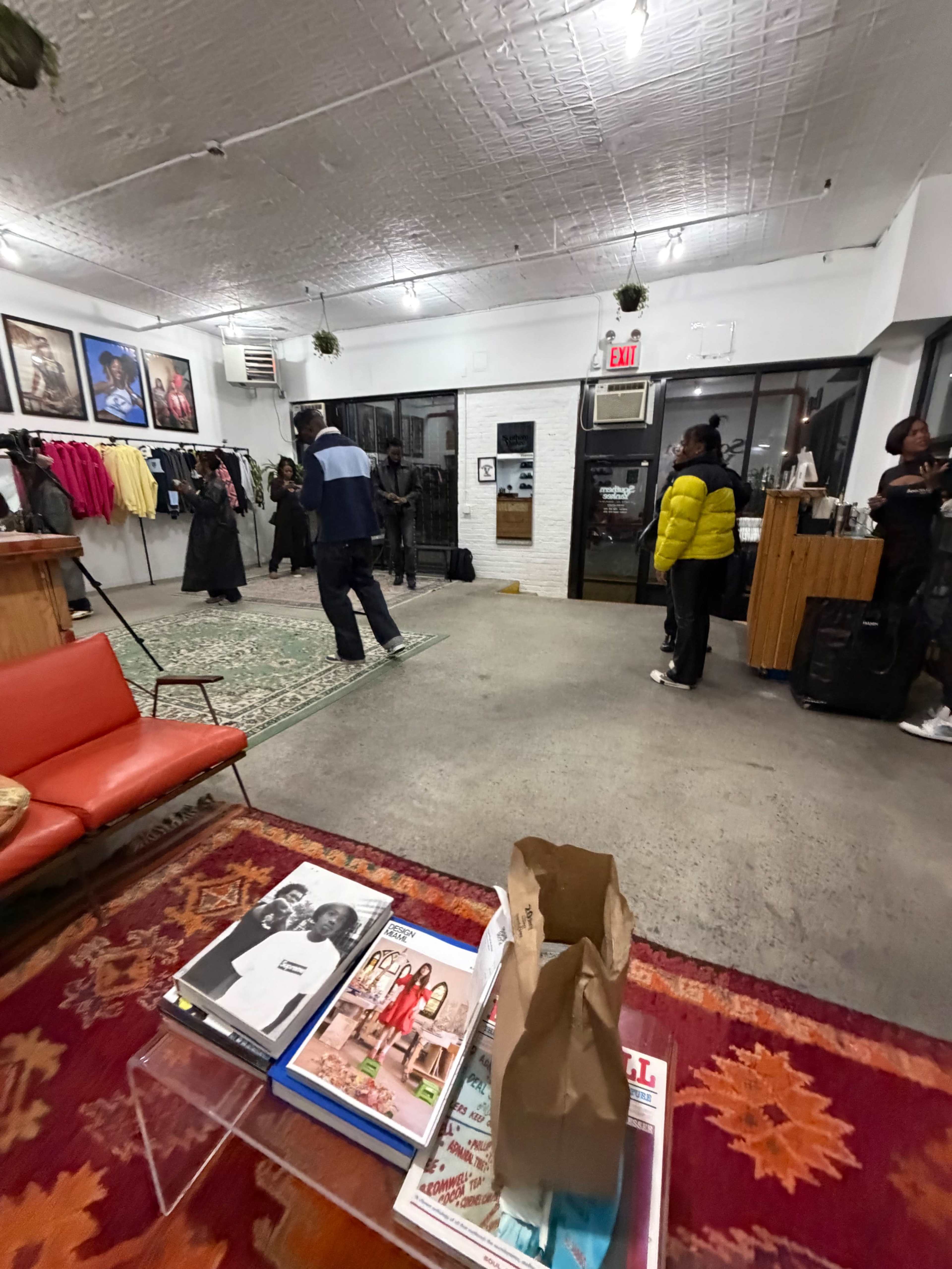 The image shows a clothing store interior with a red couch, a coffee table displaying magazines, and people browsing and interacting in a spacious, well-lit environment.