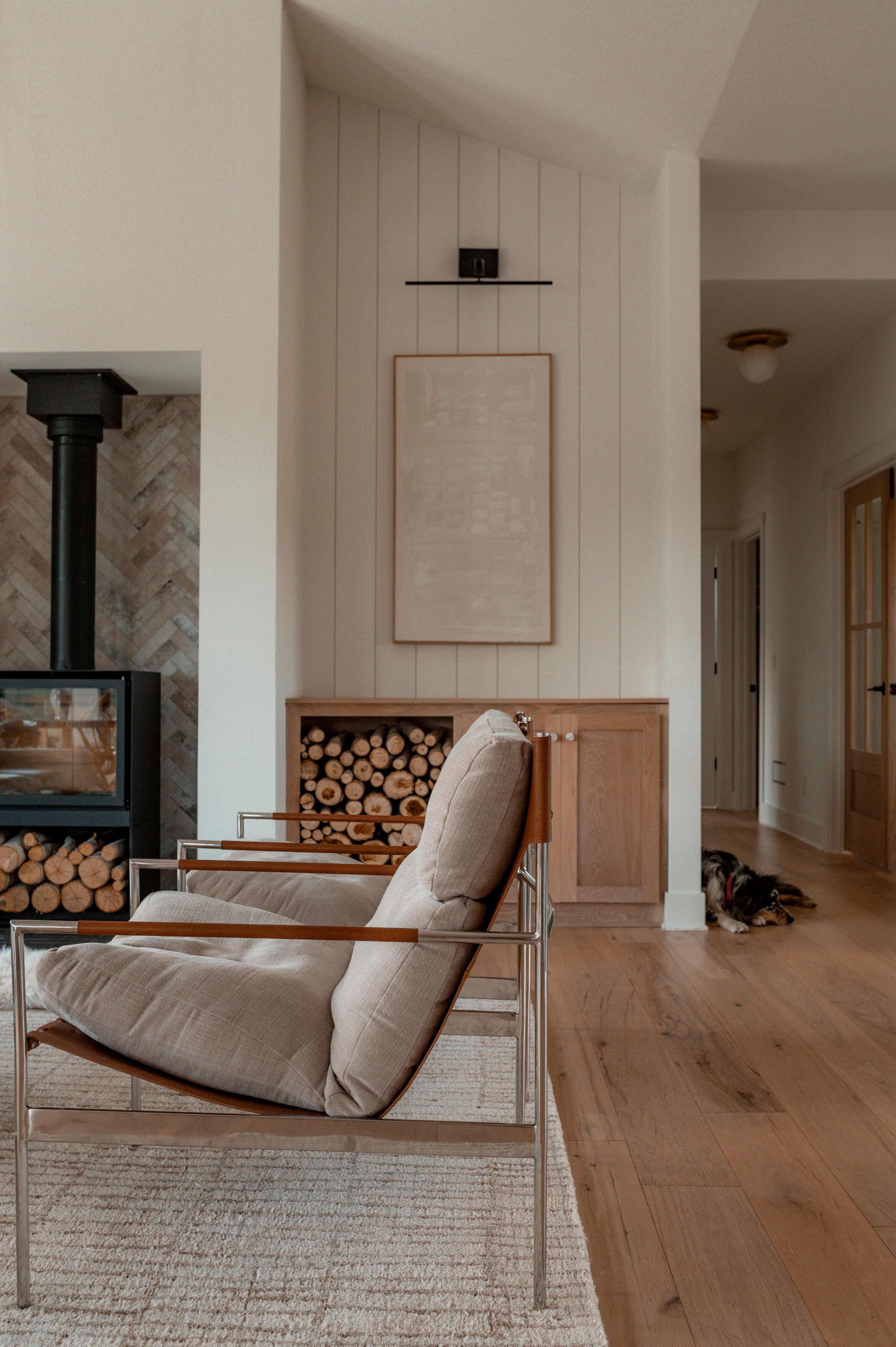 The image shows a modern living room featuring two sleek chairs, a fireplace with stacked firewood, and a wooden doorway leading to another space.