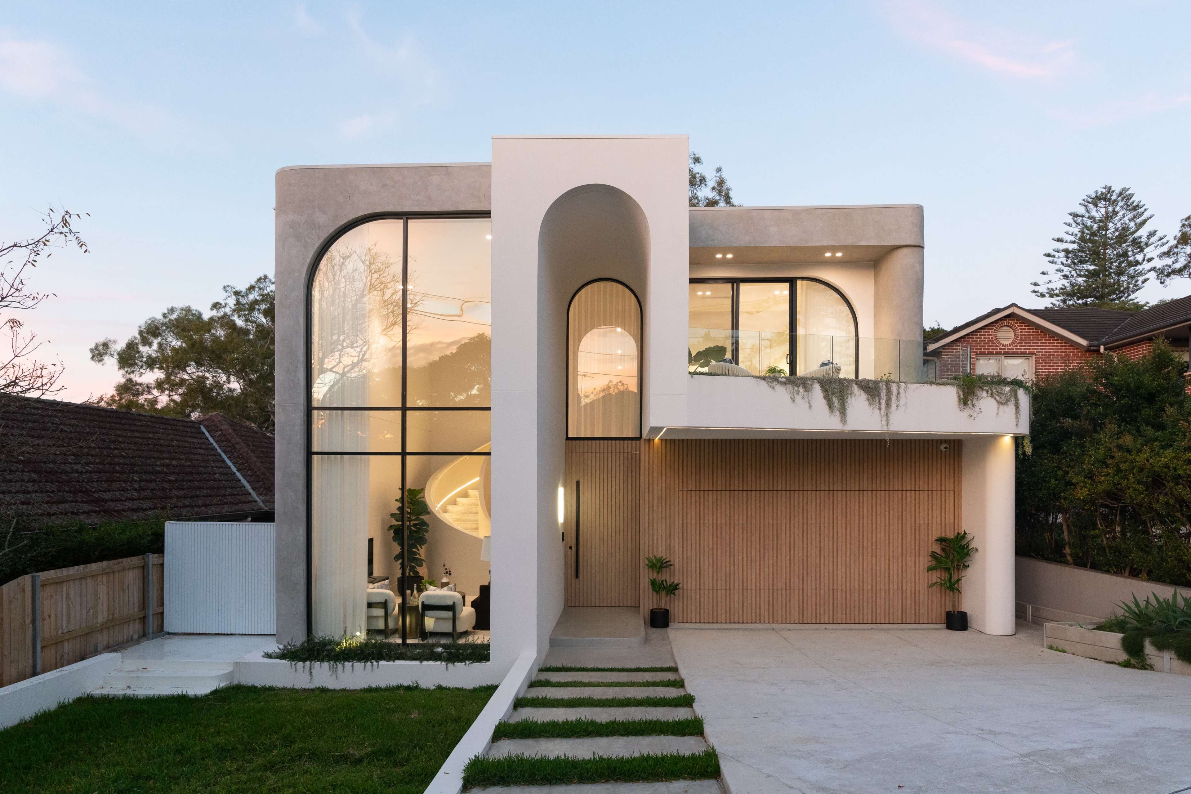 The image shows a modern residential building with large glass windows and a curved entrance, surrounded by a landscaped yard and driveway.