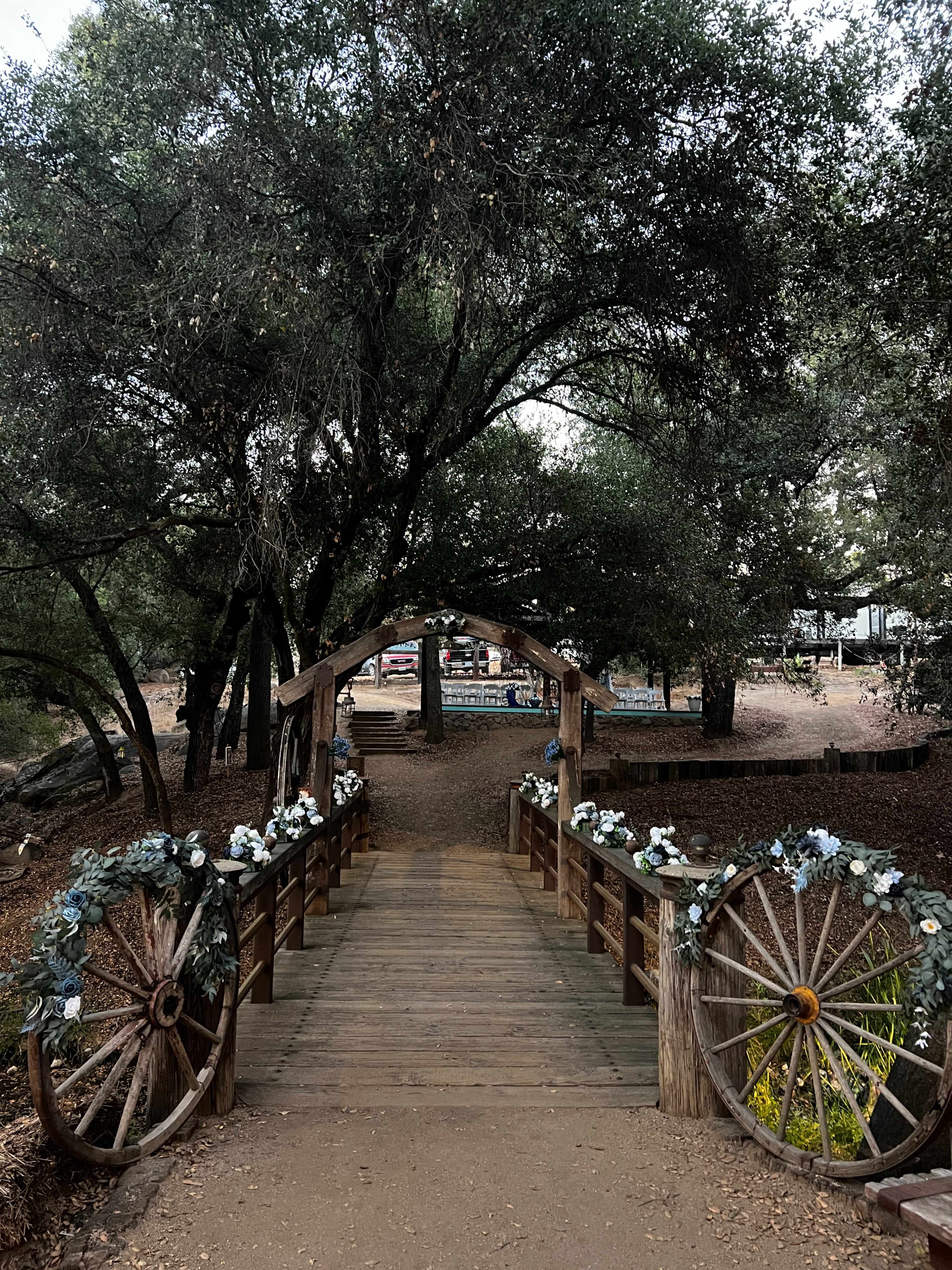 A wooden bridge adorned with floral arrangements leads through a tree-lined path in a park setting.