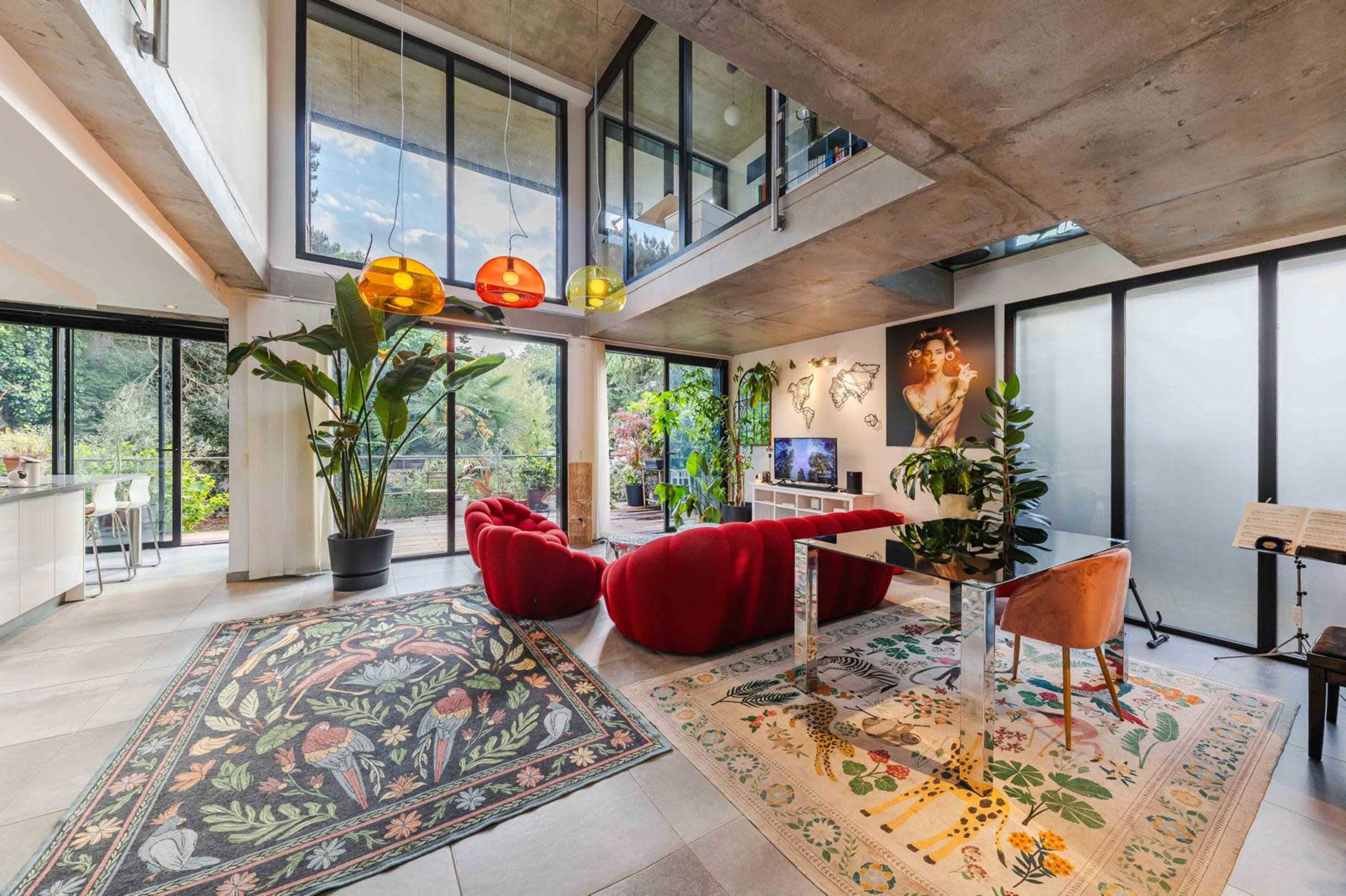 The image shows a modern, open-concept living space featuring large windows, a mix of colorful pendant lights, vibrant rugs, and contemporary furnishings, with plant decor and artwork on the walls.