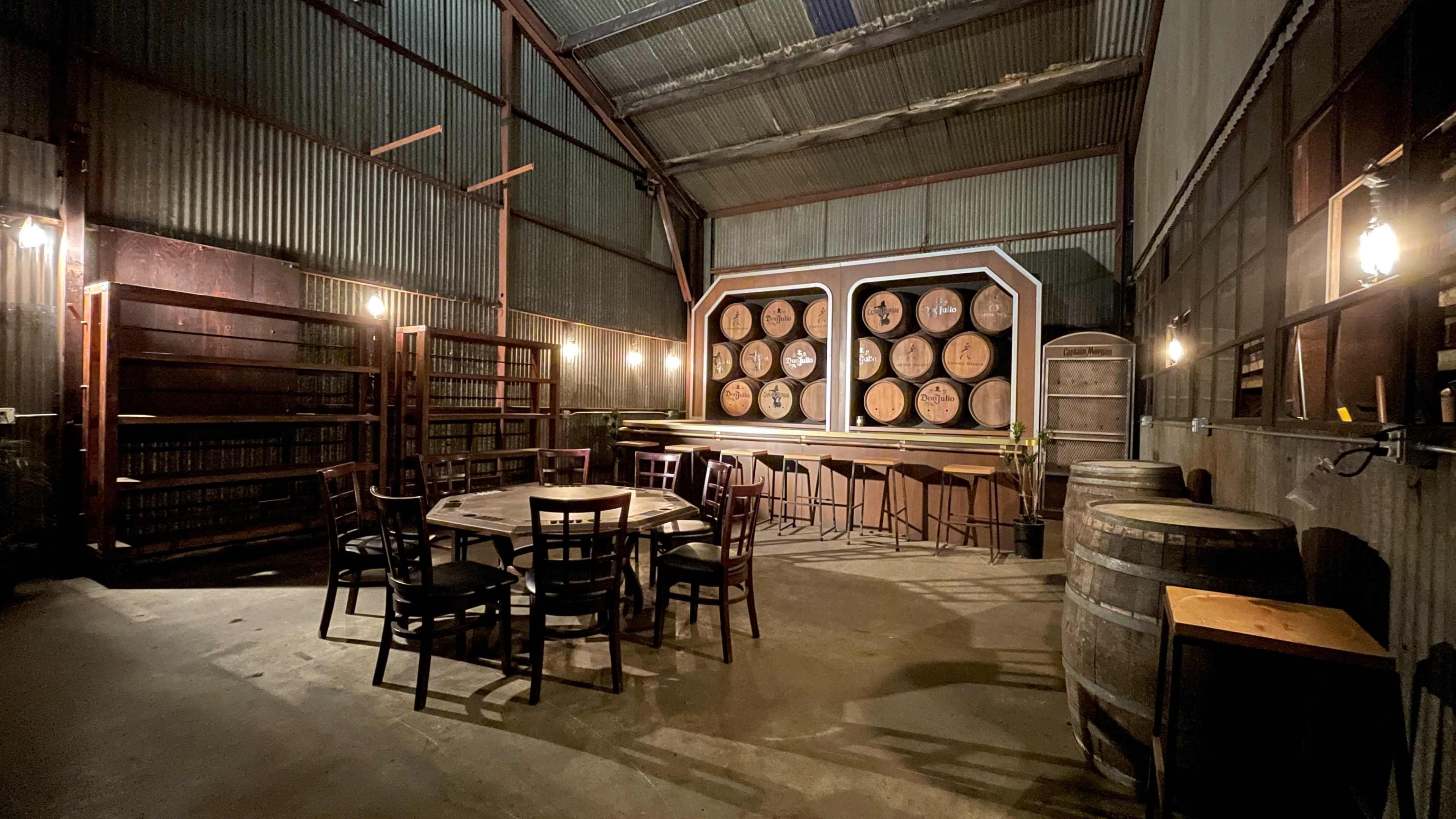 The image shows an industrial-style room featuring wooden barrels, a circular bar with large barrels behind it, and a set of tables and chairs arranged in the center.