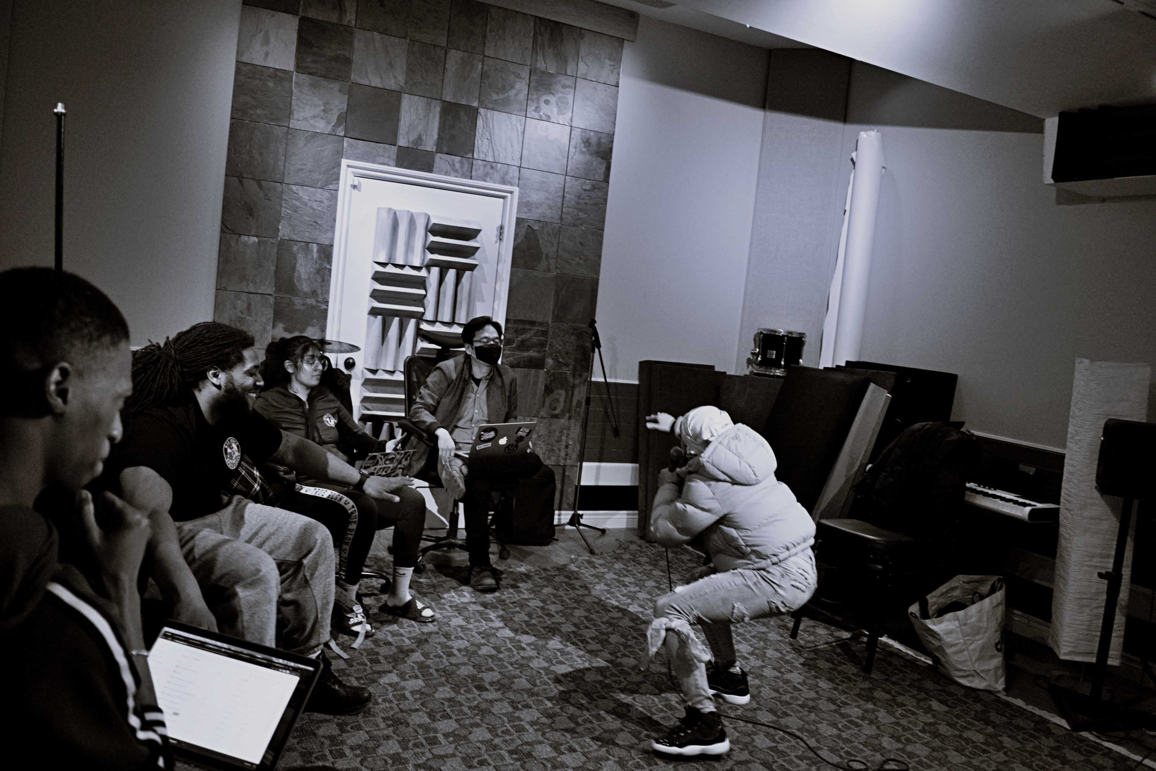 A group of six individuals watches a performer in a recording studio setting.
