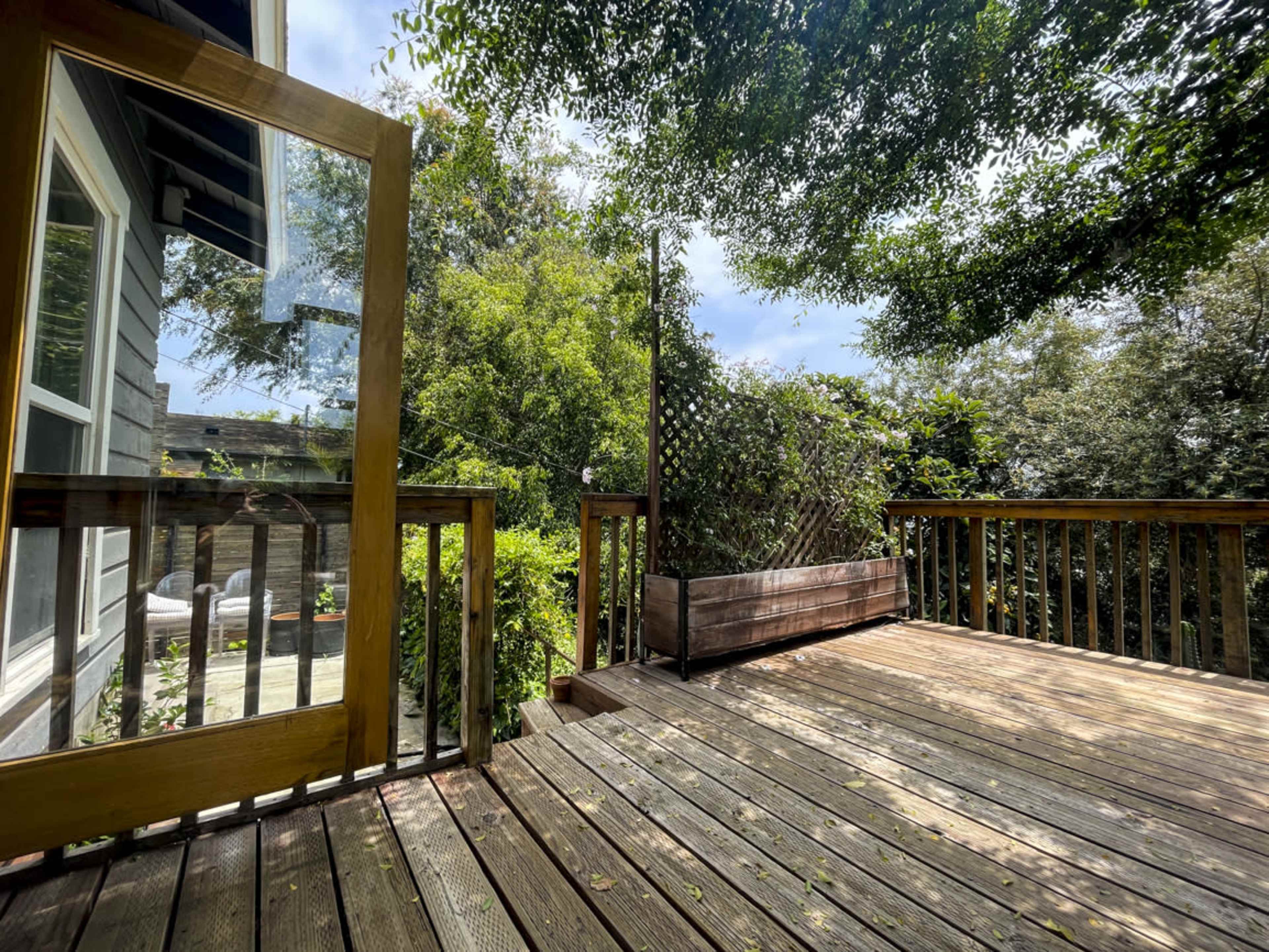 A wooden deck with an open door leads to a lush, green backyard surrounded by trees and plants.