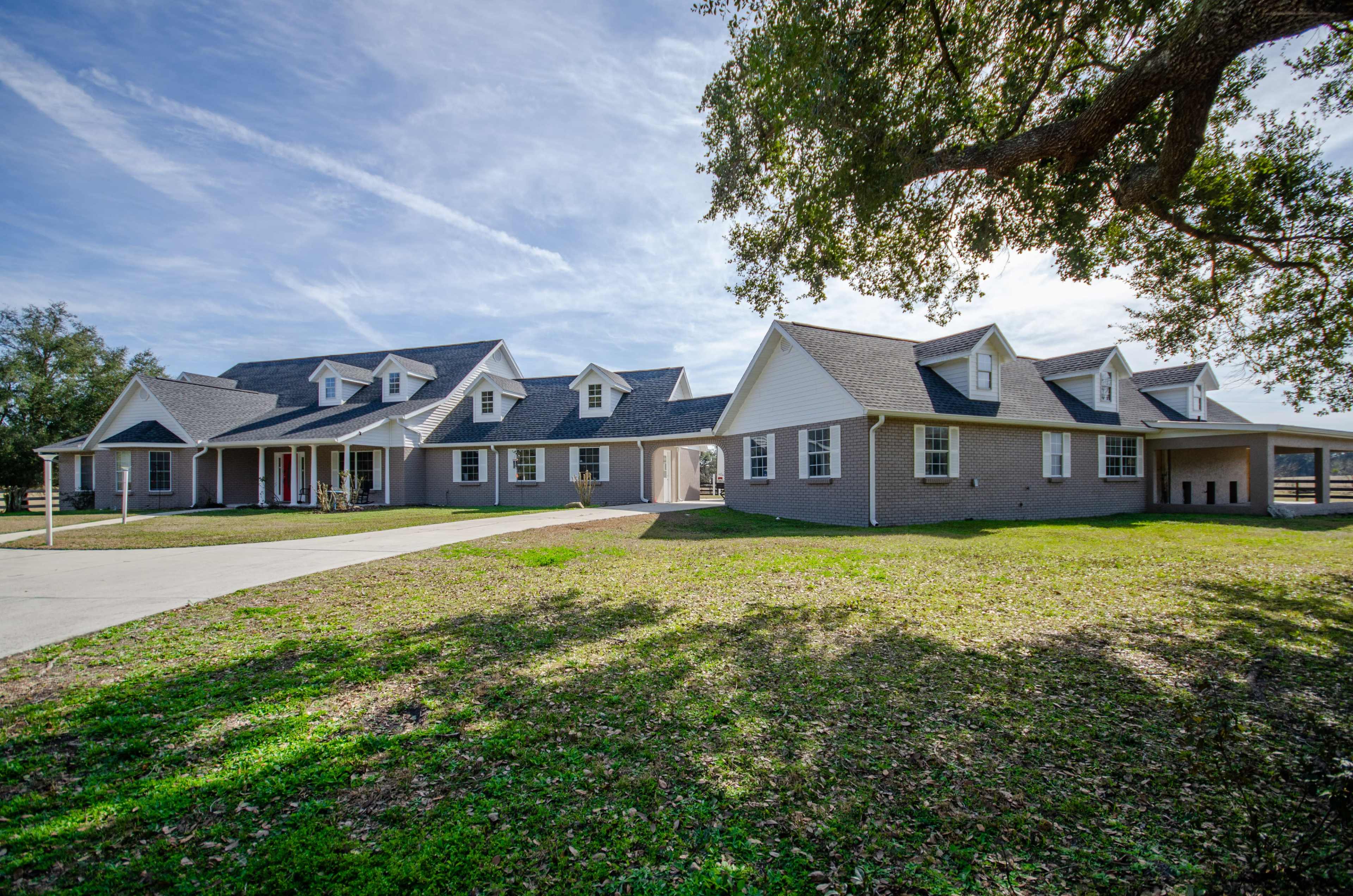 A large, two-story house with multiple gabled roofs is set on a grassy area, surrounded by trees under a blue sky.