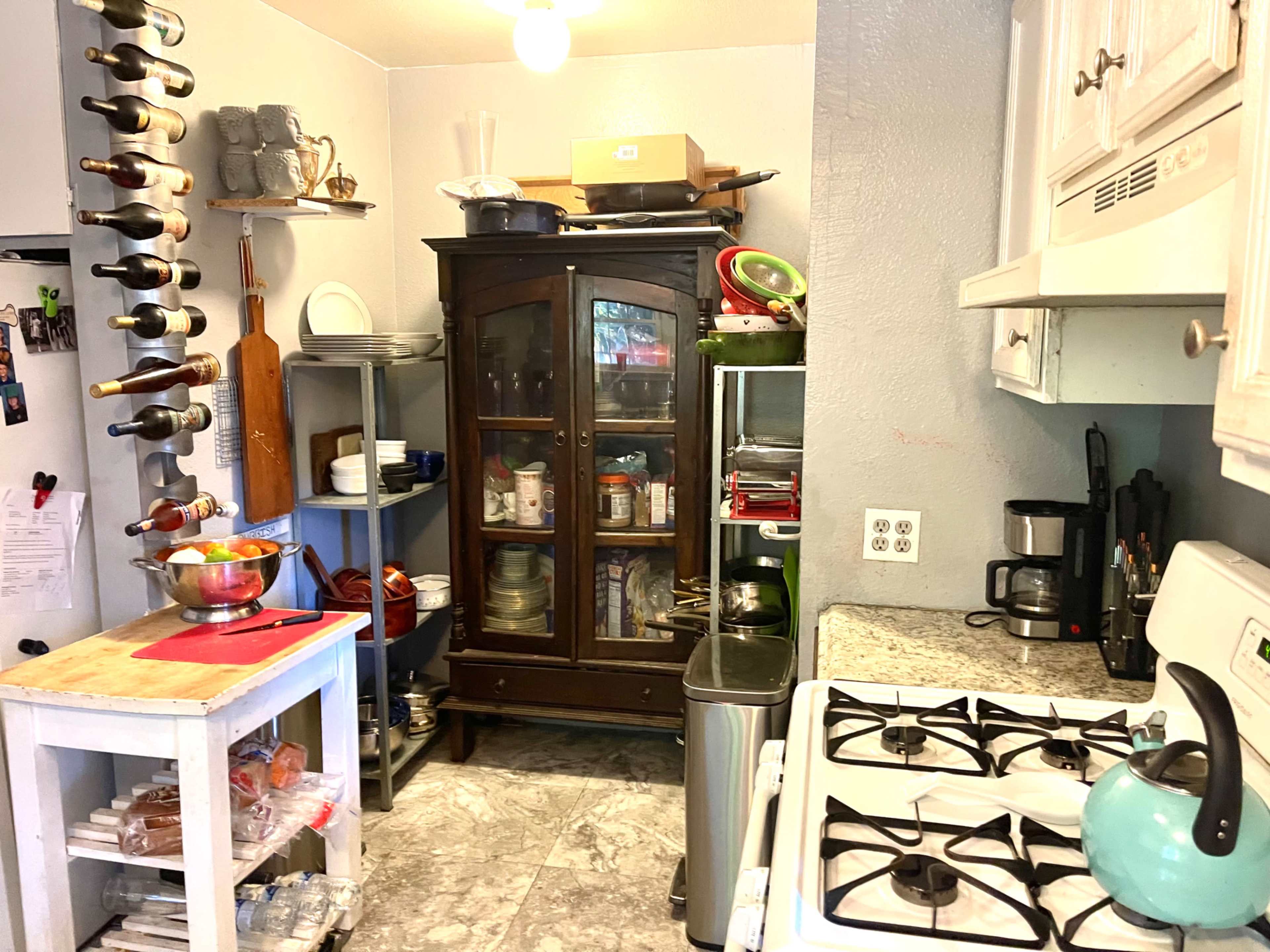The kitchen features a gray wall, a wooden dining table with a bowl of fruit, a glass-front cabinet filled with dishes, and a stove beside various kitchen appliances and a wine rack.