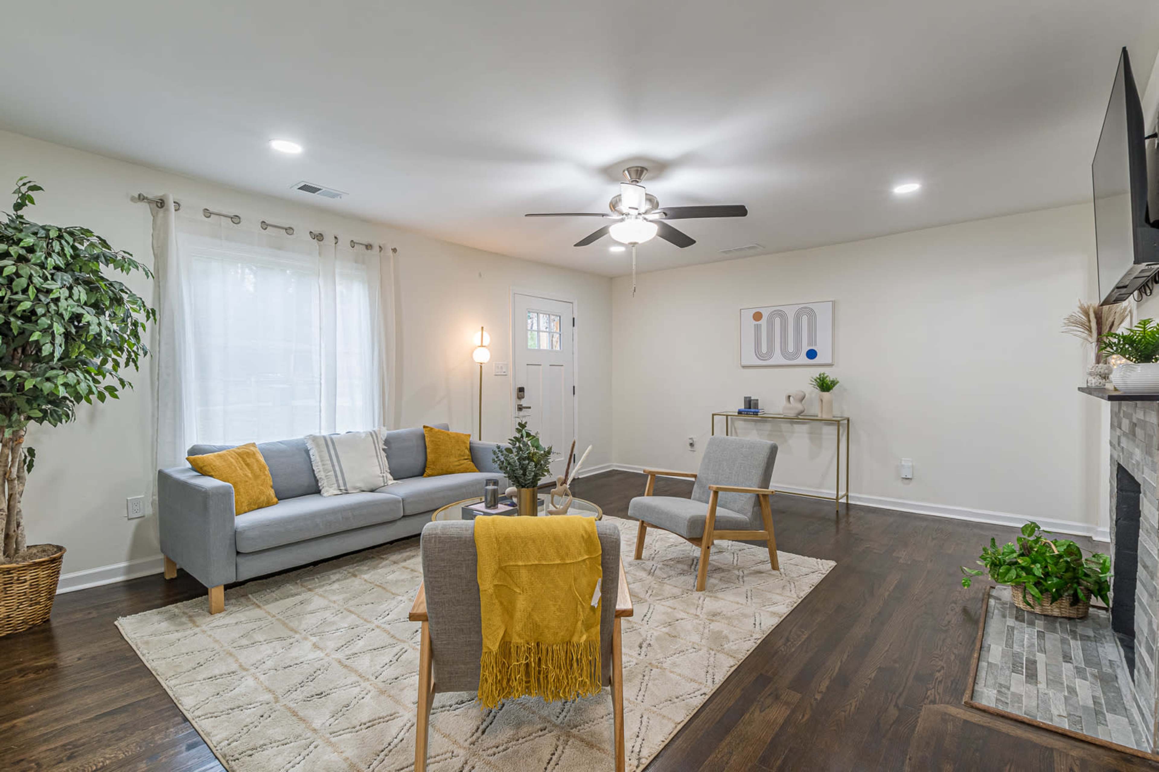 A modern living room features a grey couch, wooden chairs, a coffee table, and a large area rug, with a ceiling fan, light, and plants in a bright space.