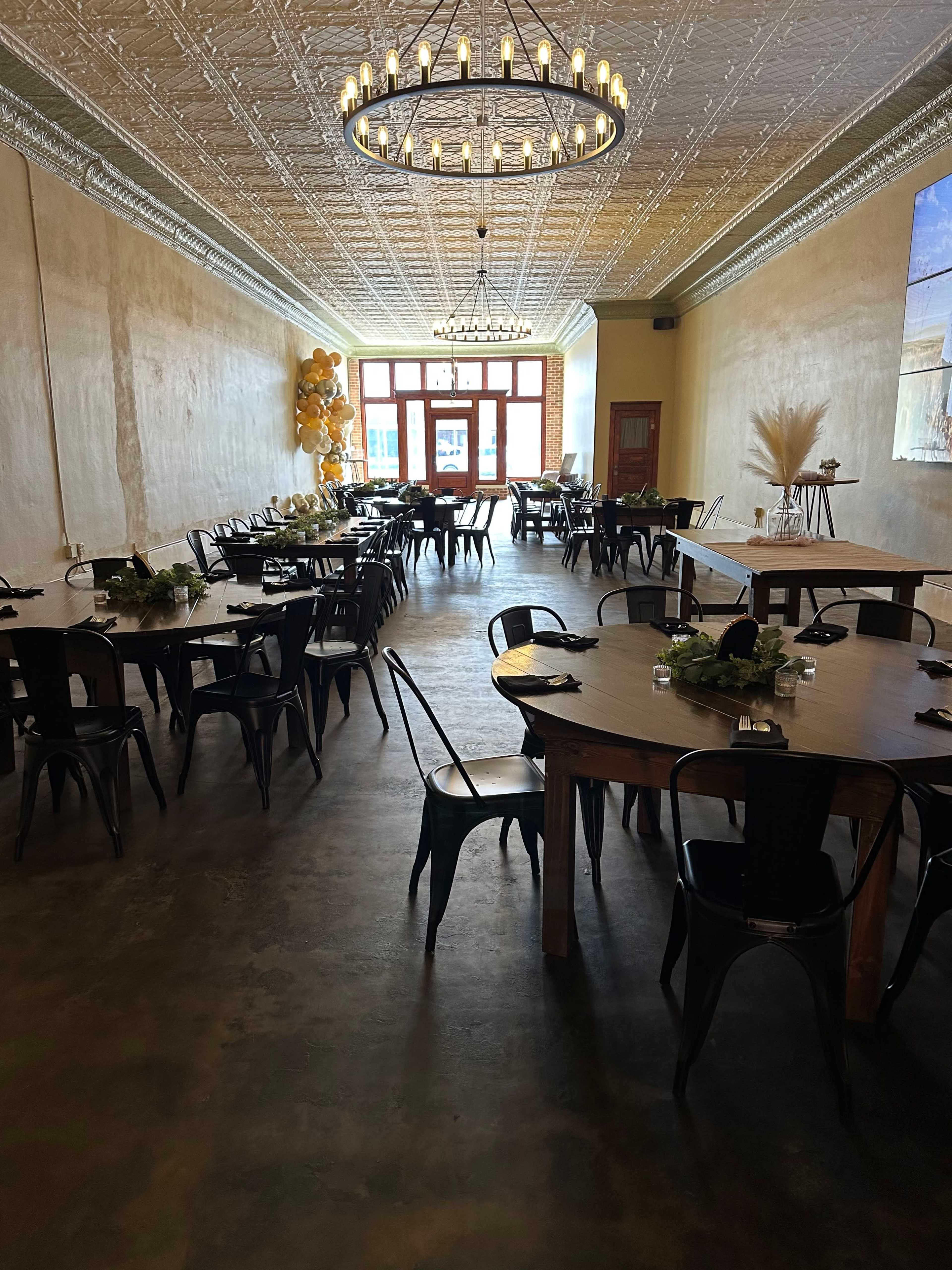 The image displays a spacious restaurant interior featuring round and rectangular tables set with black chairs, with decorations and a large window at one end.
