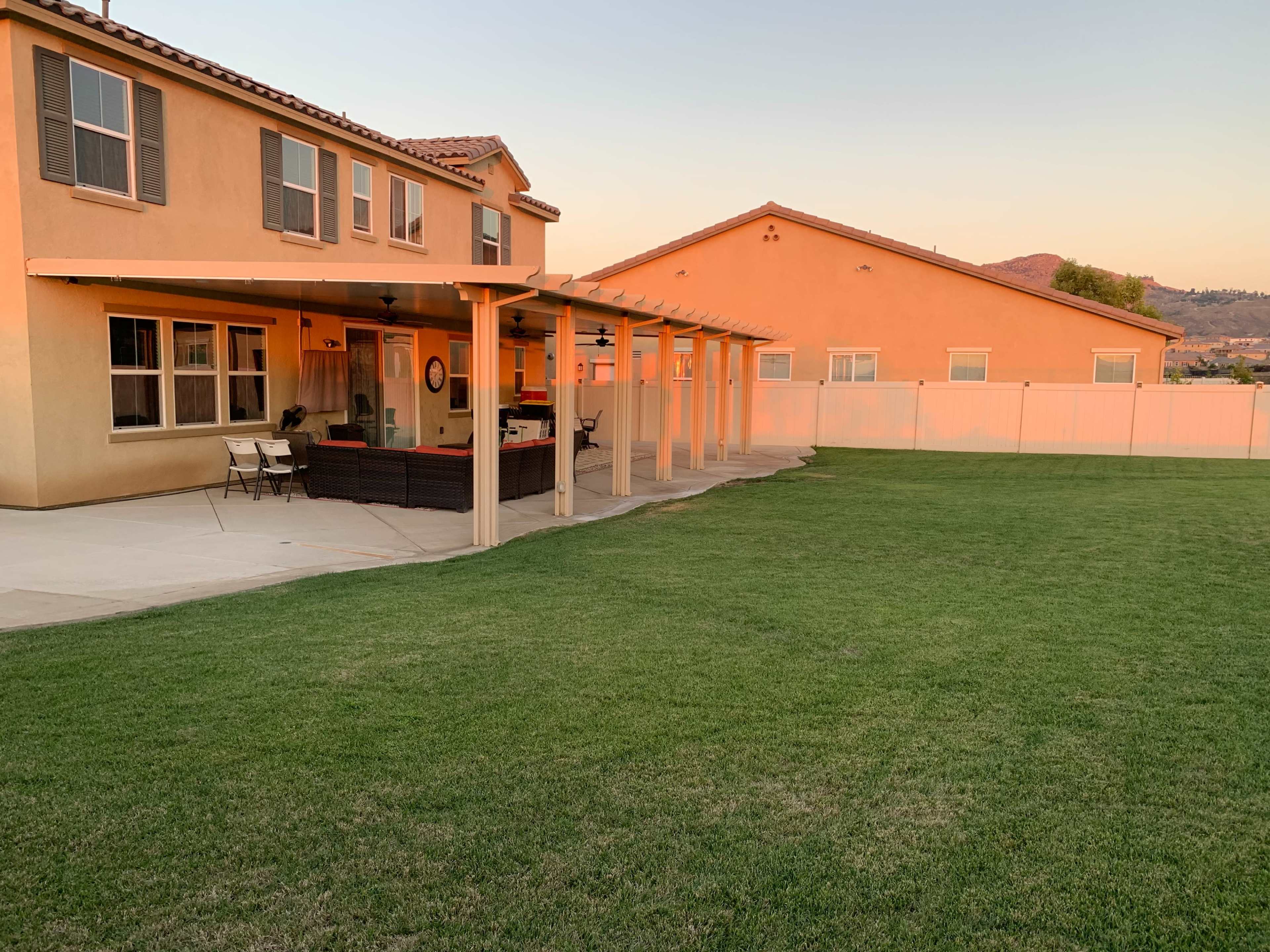 A backyard with a patio covered by a pergola, adjacent to two houses and a grassy lawn.