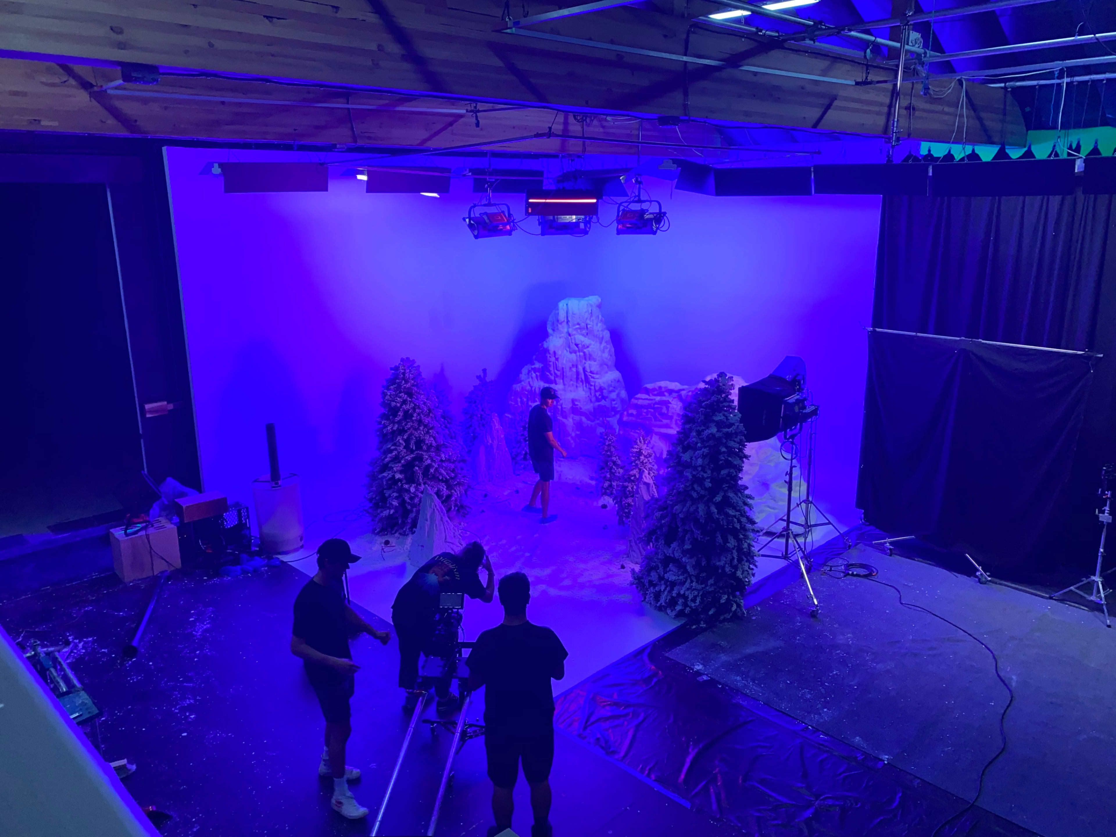 A film crew sets up a winter-themed backdrop featuring artificial snow and pine trees in a studio illuminated by blue lights.