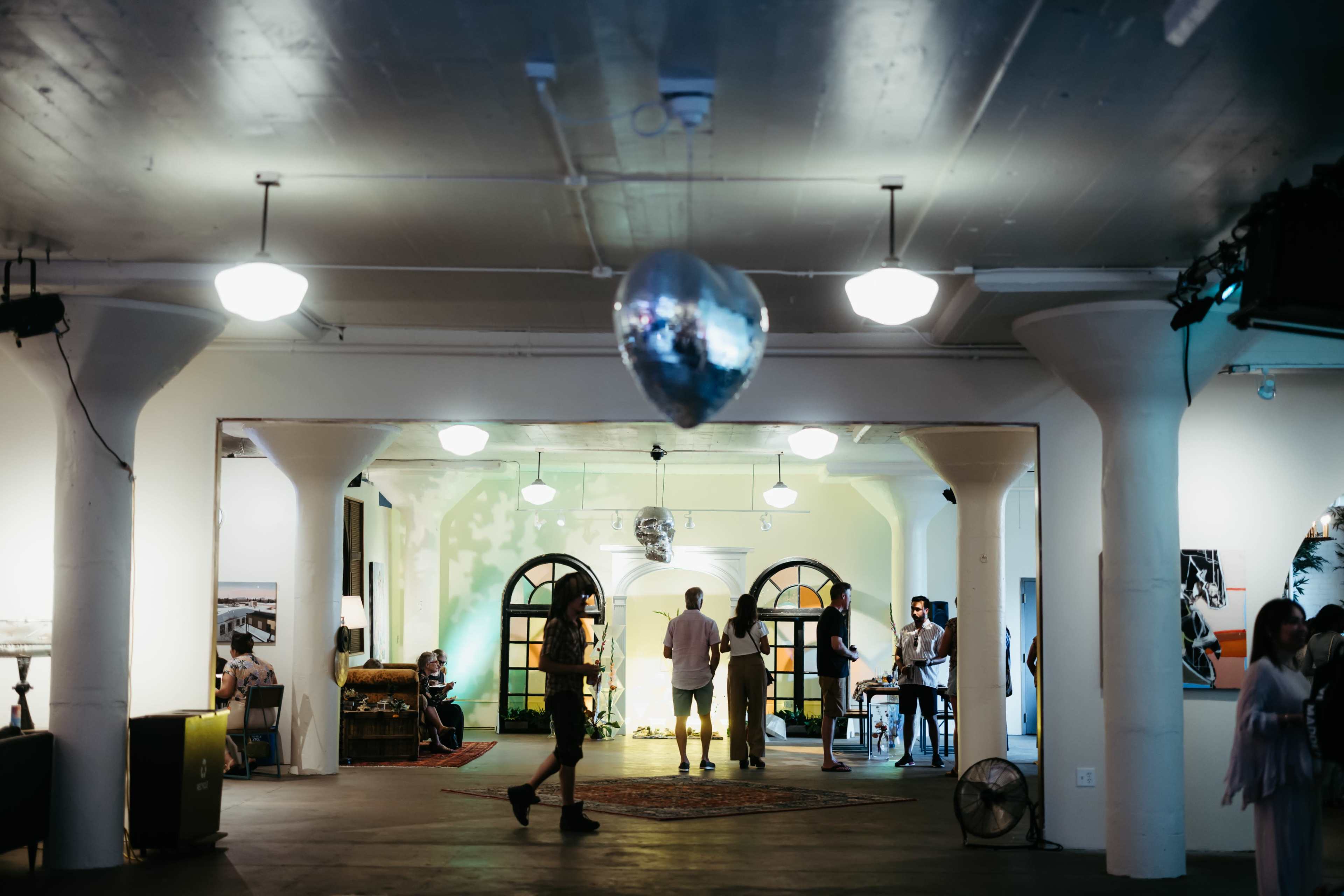 An interior space with polished floors, pillars, and various people engaged in conversation under soft lighting, including a disco ball hanging from the ceiling.