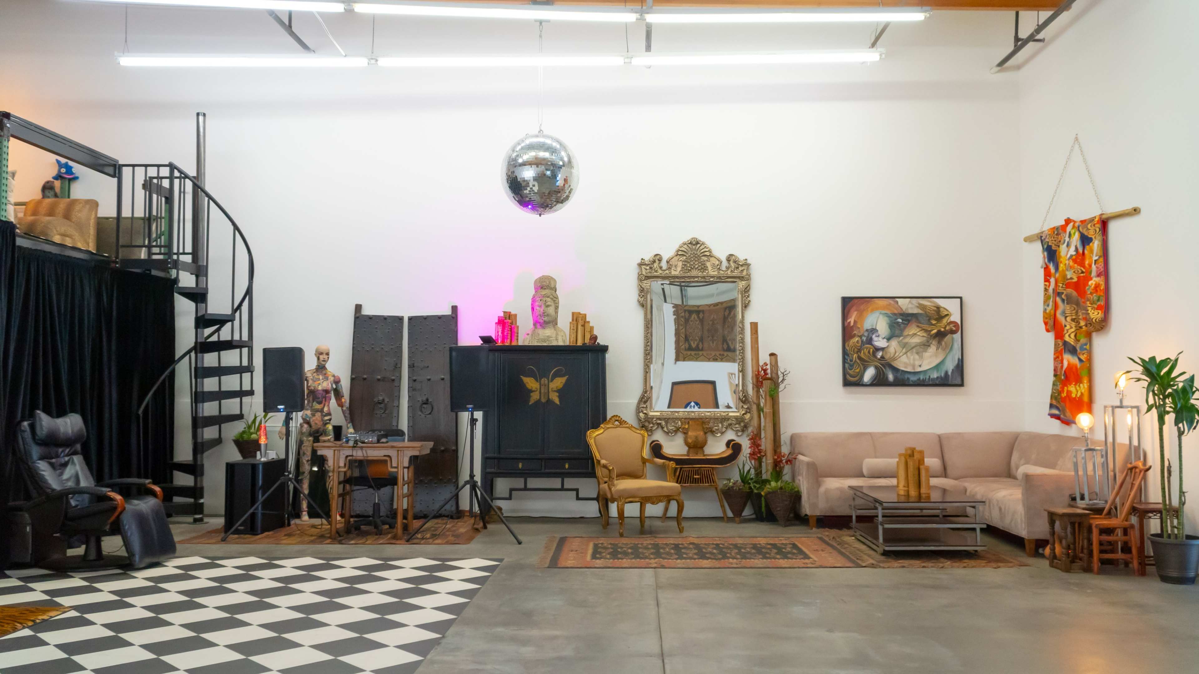 The image shows an industrial-style studio space featuring a spiral staircase, various furniture pieces, a disco ball, and decorative art on the walls.