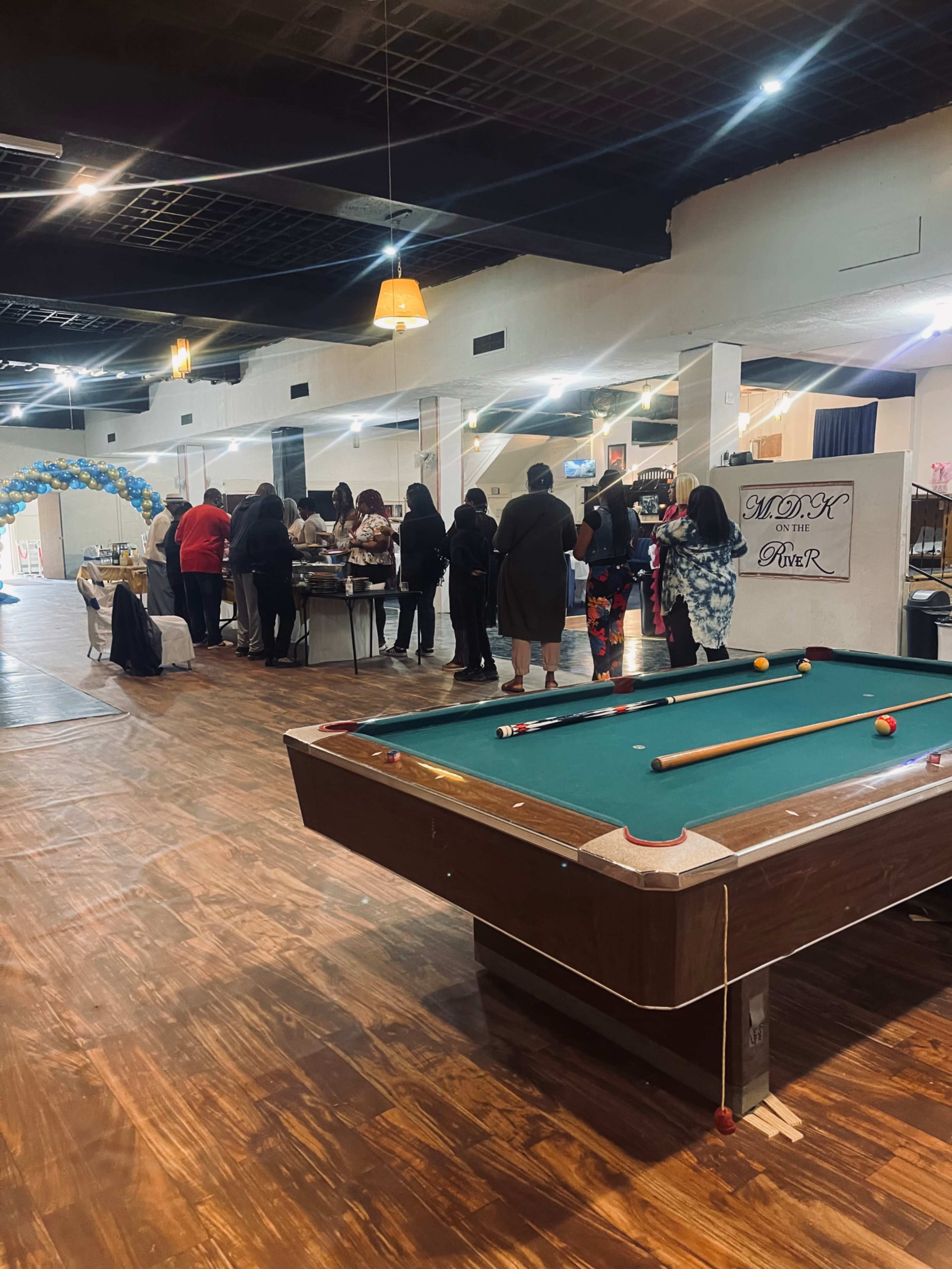 The image shows a large indoor space with a pool table in the foreground and a group of people gathered around tables in the background.