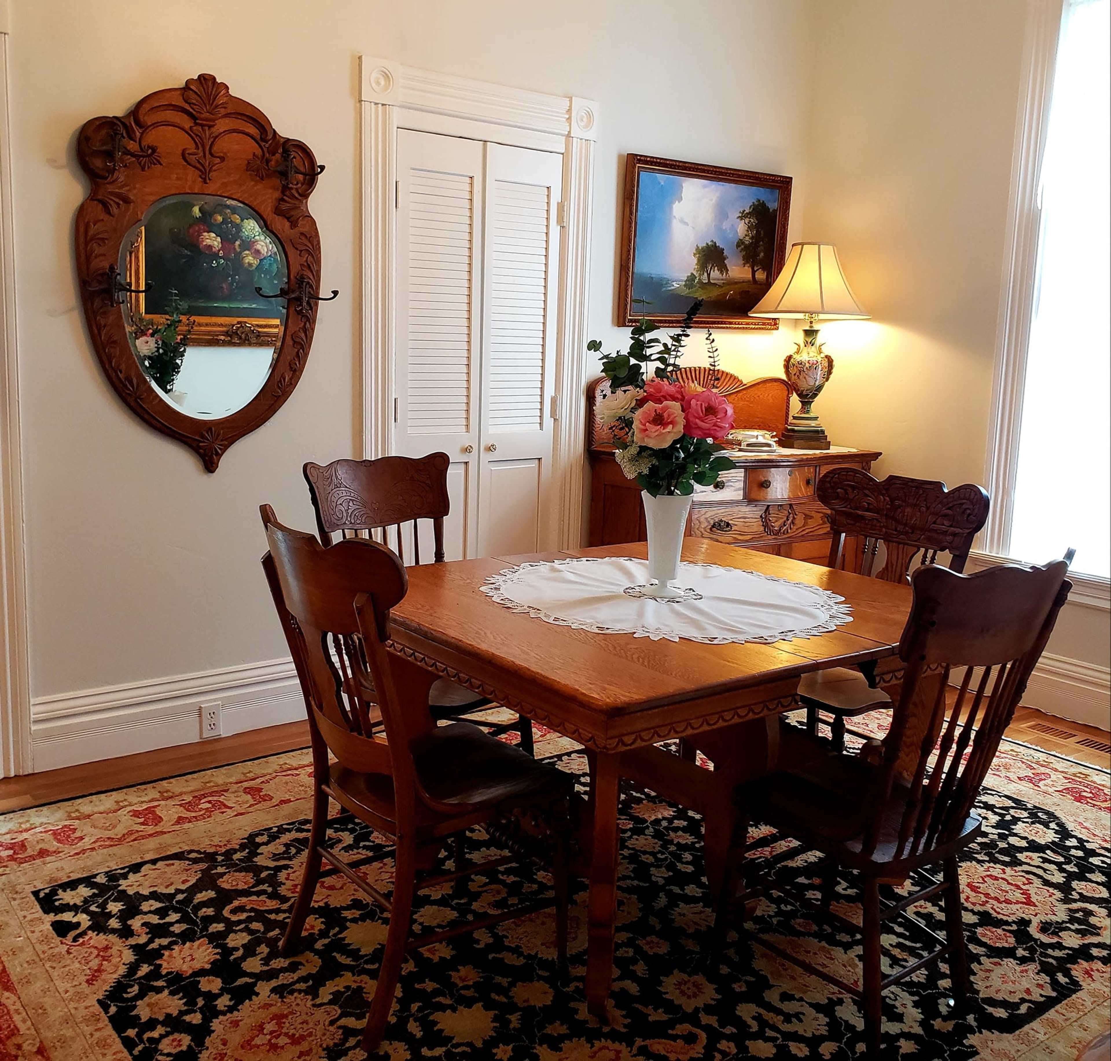 A wooden dining table surrounded by four chairs is set in a room with a floral centerpiece, a mirror on the wall, and a lamp beside a framed painting.