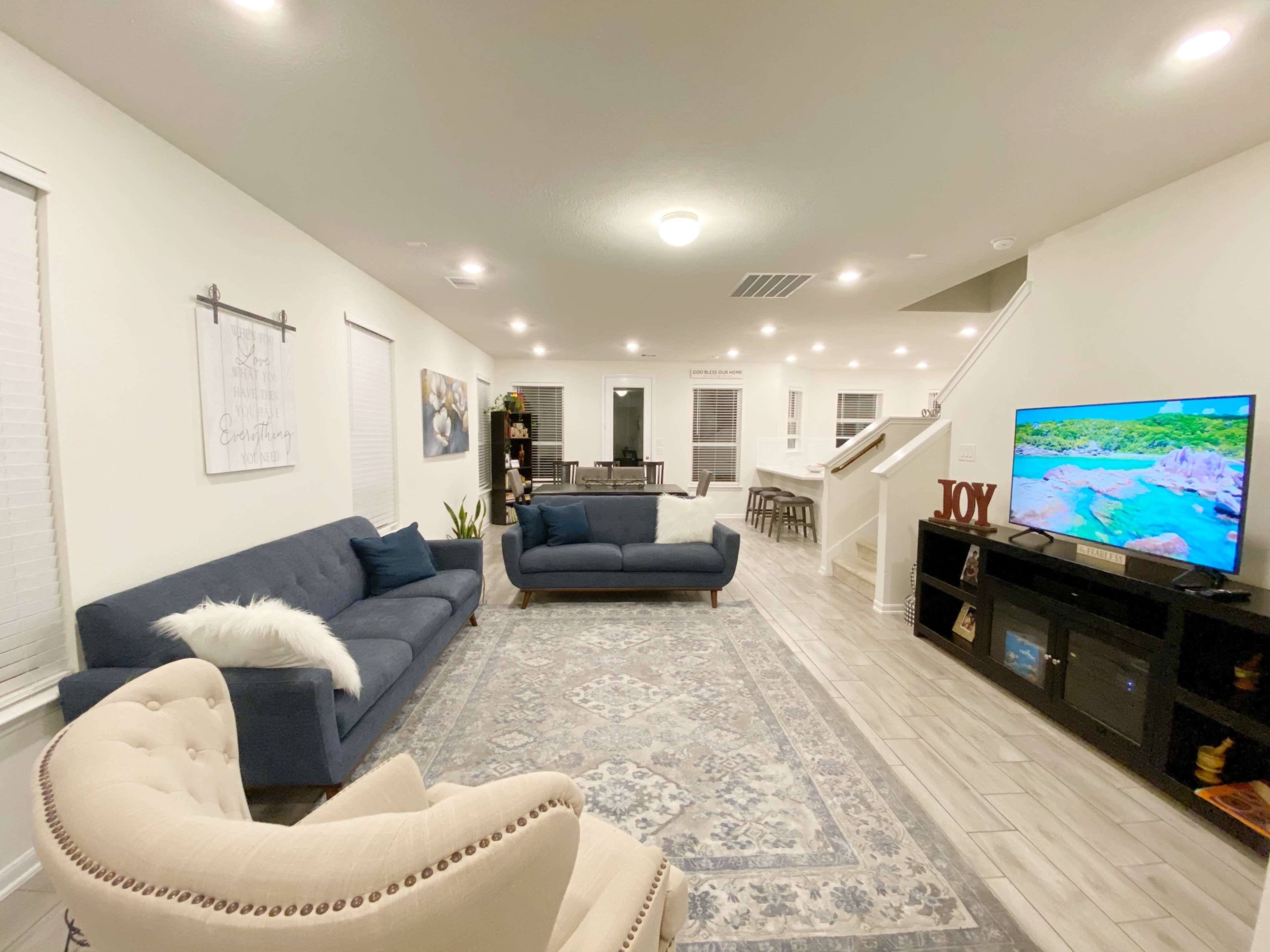 A modern living room features two blue sofas, a beige armchair, a large area rug, and a television on a black console against a light-colored wall.