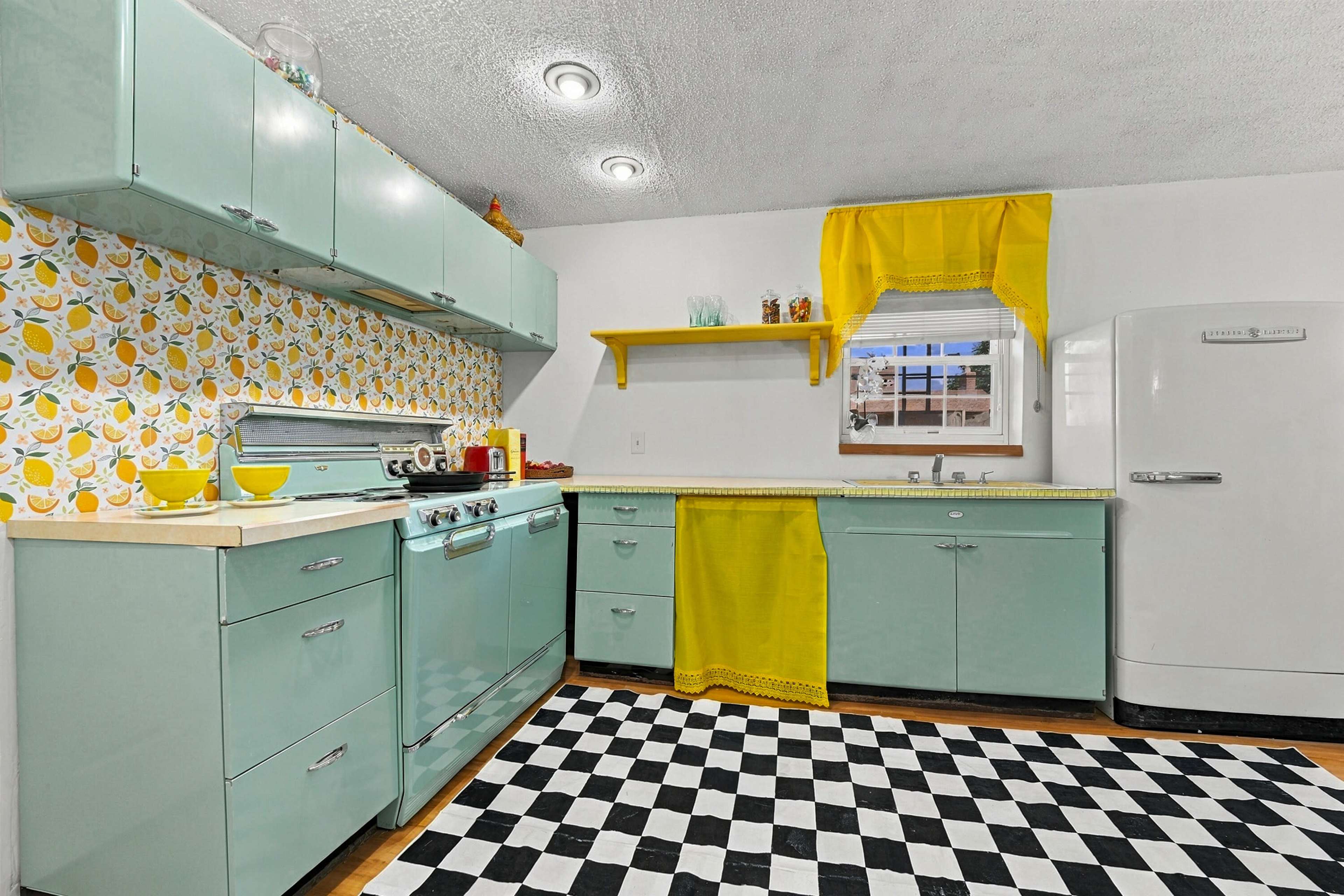 The image depicts a retro-style kitchen featuring mint green cabinets, a black and white checkered floor, and yellow accents, including a curtain and a dish towel.