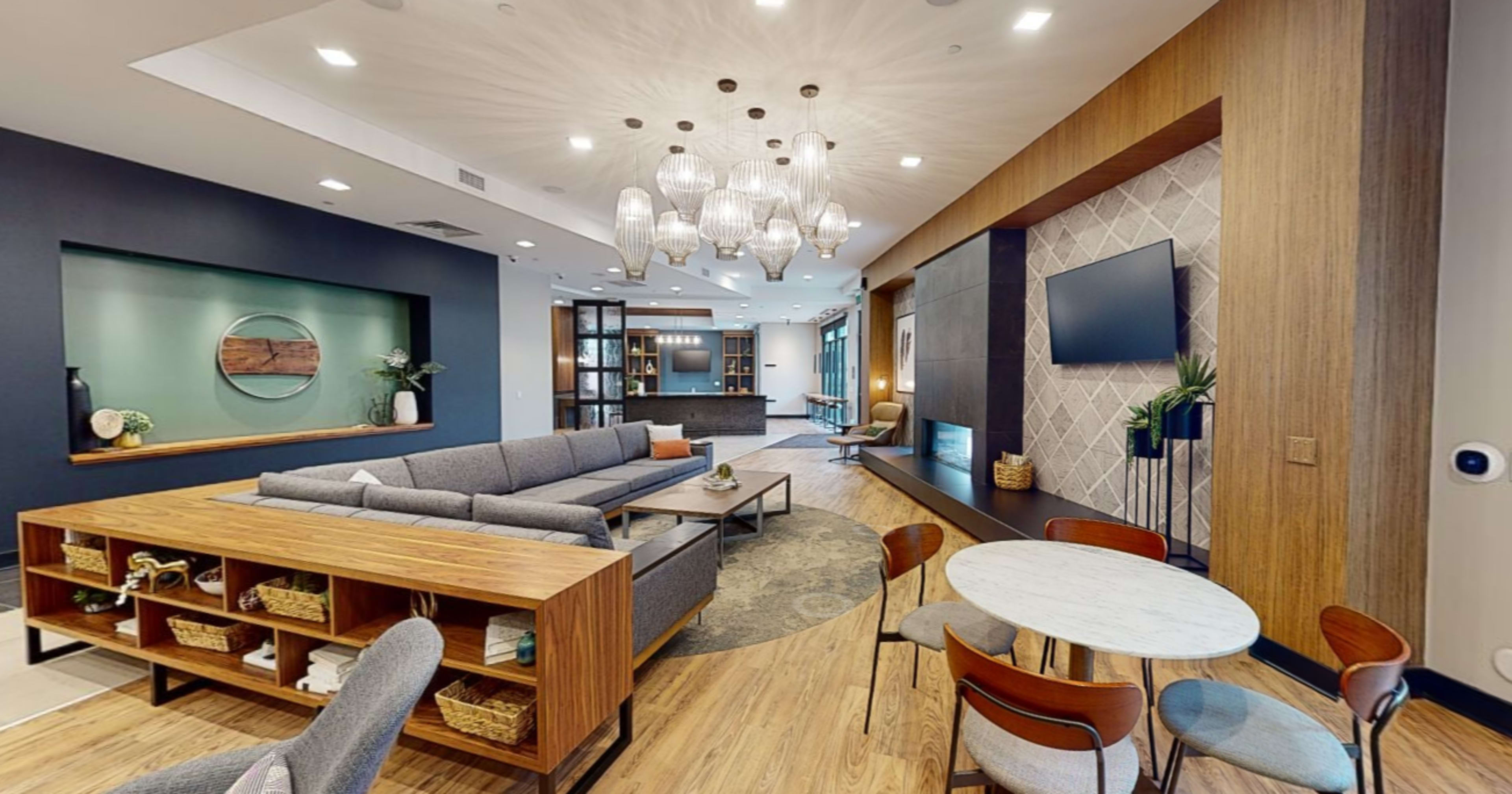 The image shows a modern lounge area featuring grey sofas, a wooden table, a round marble dining table, and large light fixtures above, with a television mounted on the wall.