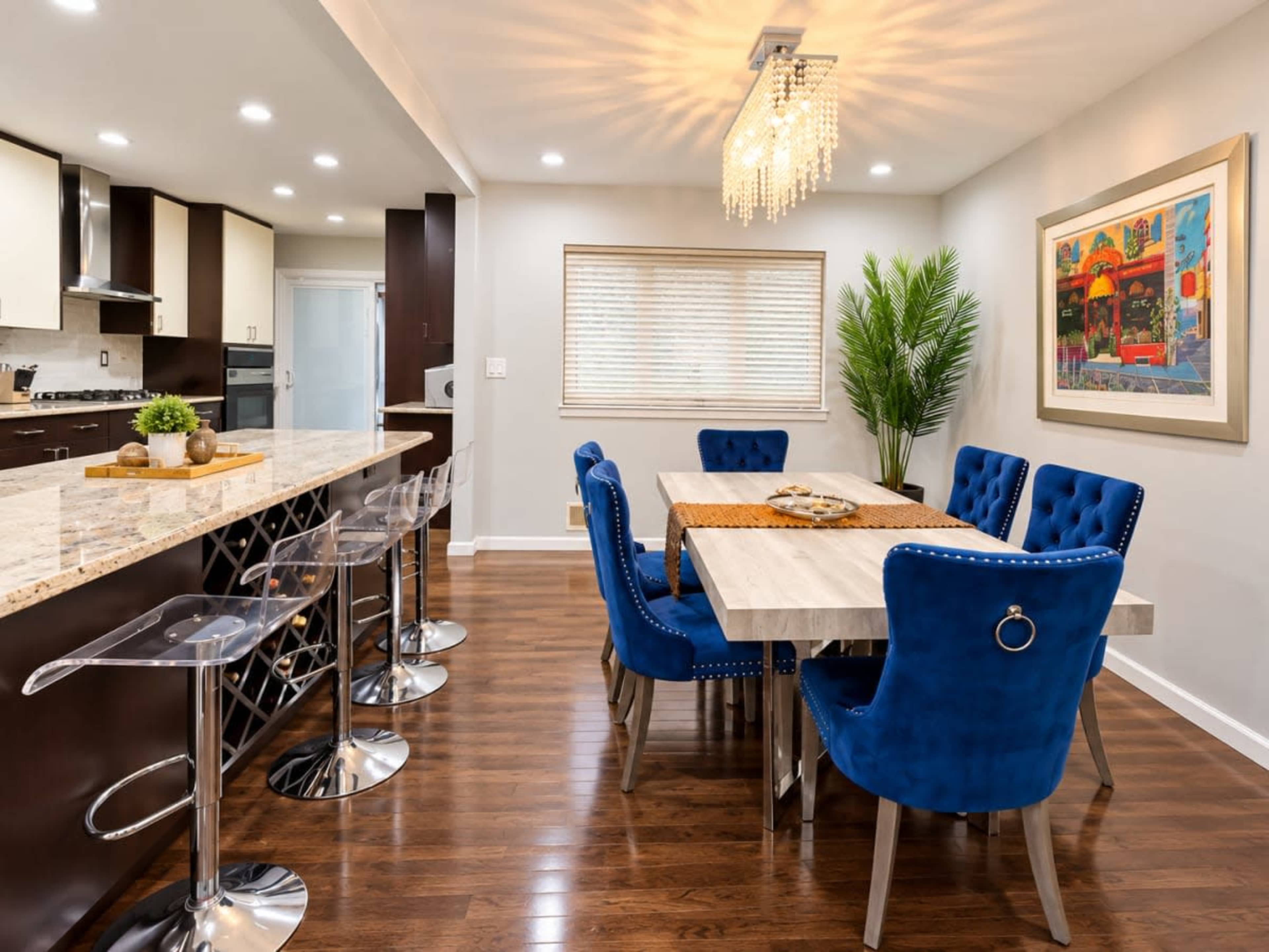 A modern kitchen and dining area features a granite countertop, a six-person dining table with blue upholstered chairs, and a decorative chandelier.