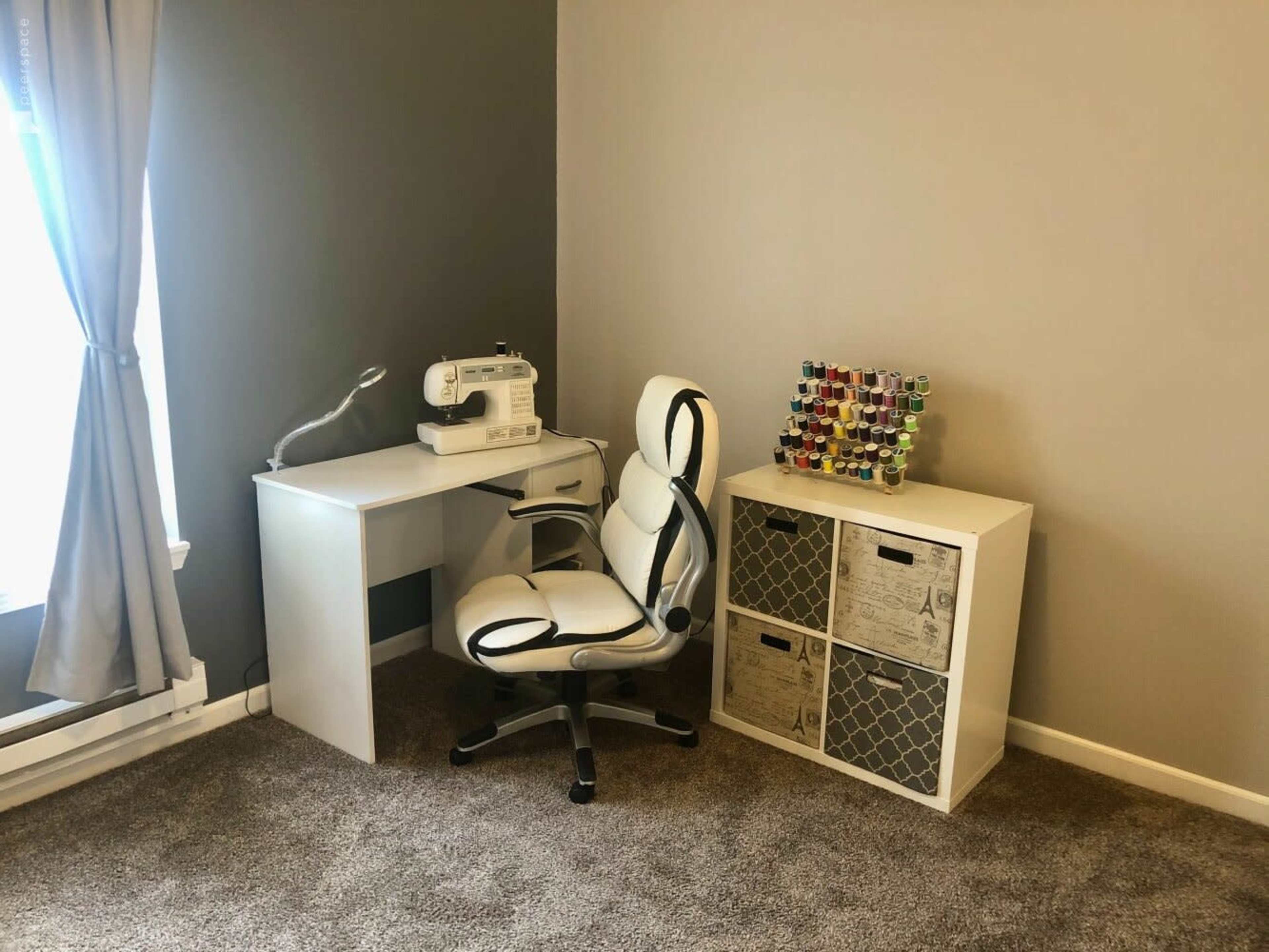 A white sewing machine is placed on a minimalist desk next to a chair and a shelving unit filled with colorful spools of thread.