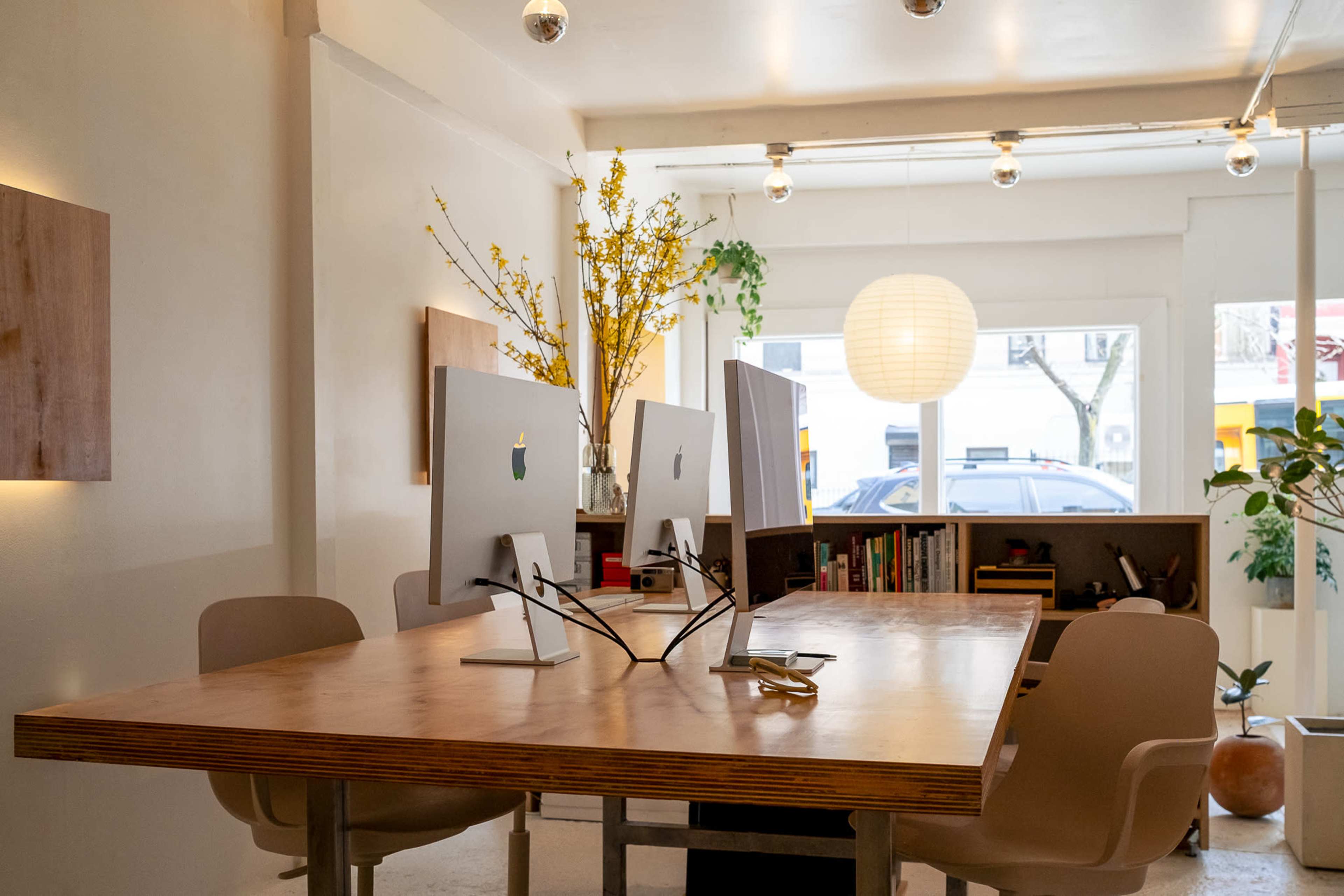The image shows a modern office space with a wooden table, two Apple monitors, and minimalist decor, including plants and a paper lantern.