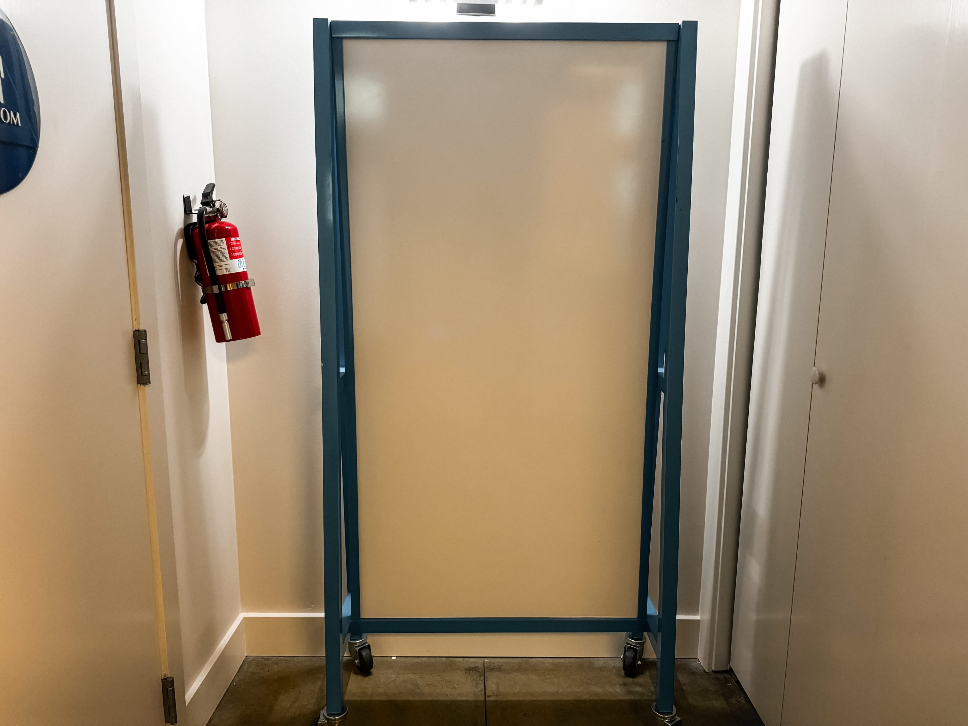 A blue mobile whiteboard stands against a white wall, next to a fire extinguisher.