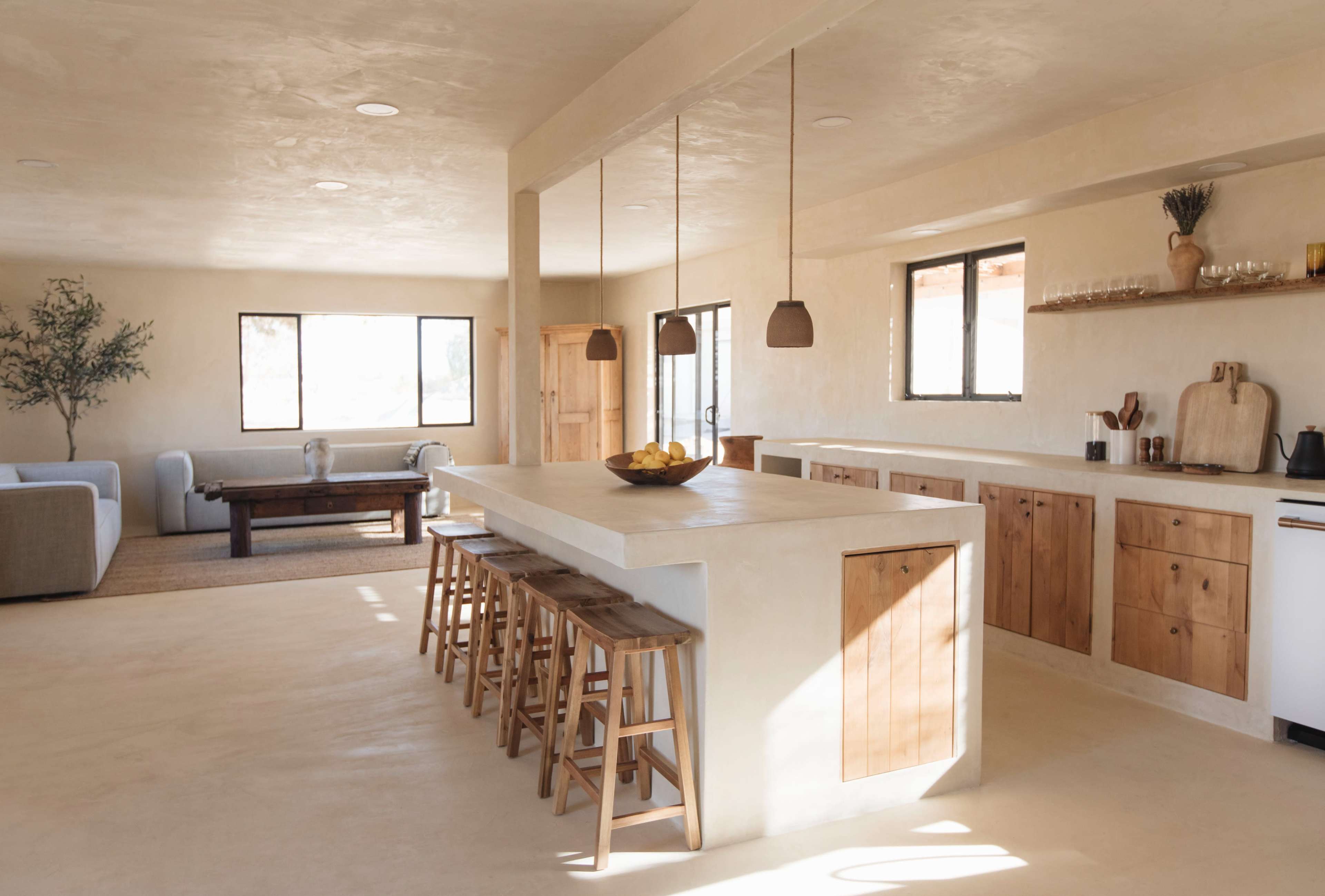 A spacious, modern kitchen features a large island with wooden stools and minimalist cabinetry, illuminated by natural light from multiple windows.