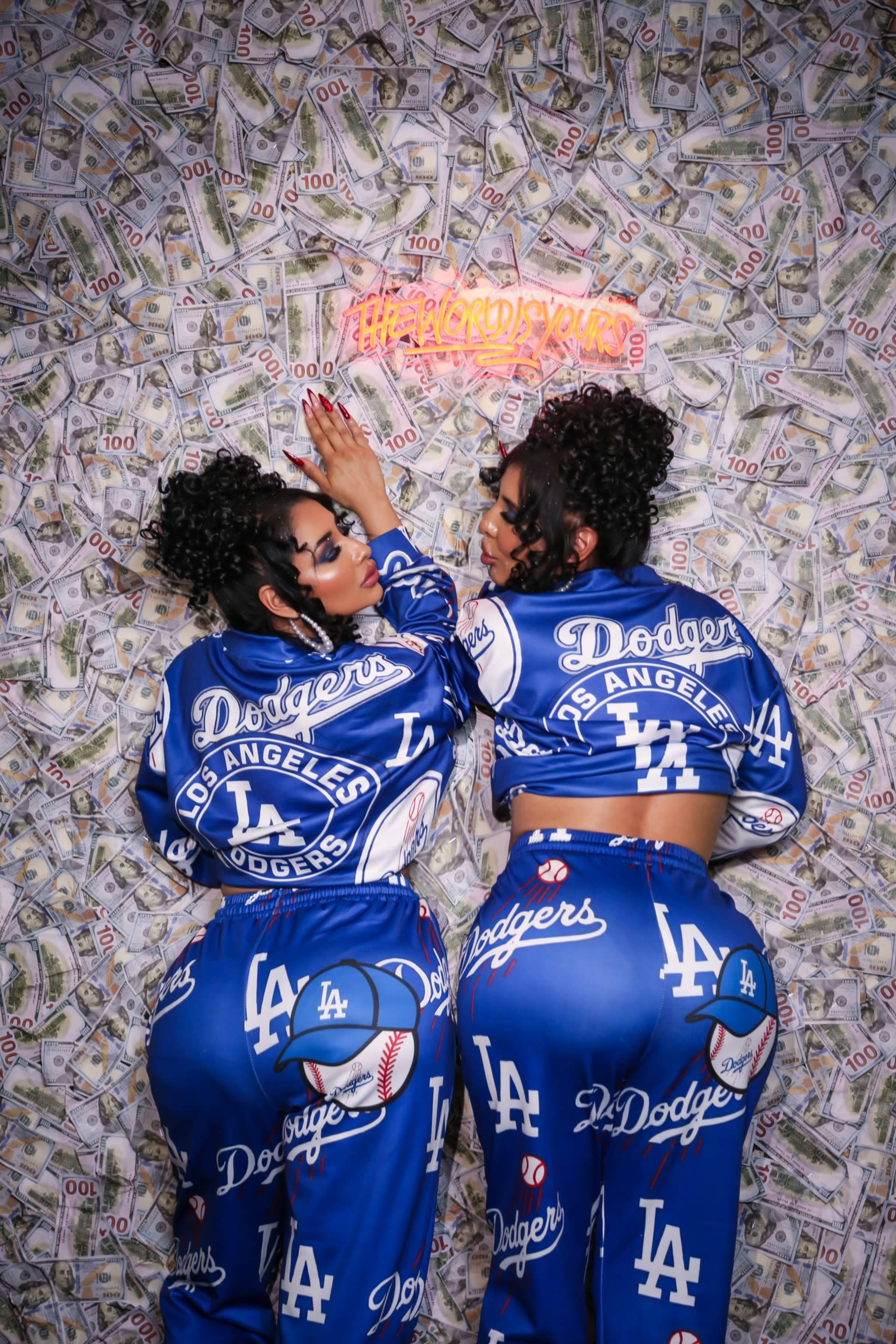 Two women in matching blue Los Angeles Dodgers outfits pose against a wall covered in images of money.