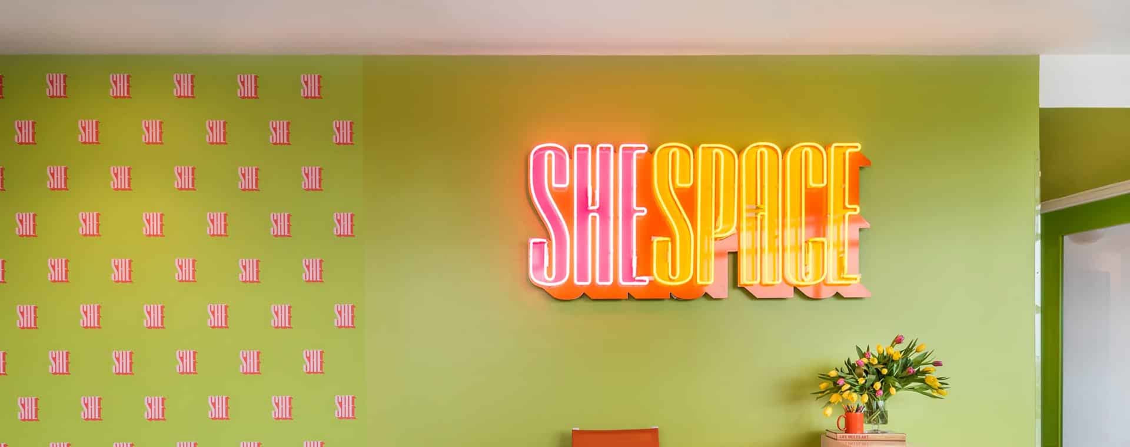 The image shows a brightly colored room with a vibrant green wall featuring a neon sign that reads "SHESPACE" and a floral arrangement on a table.