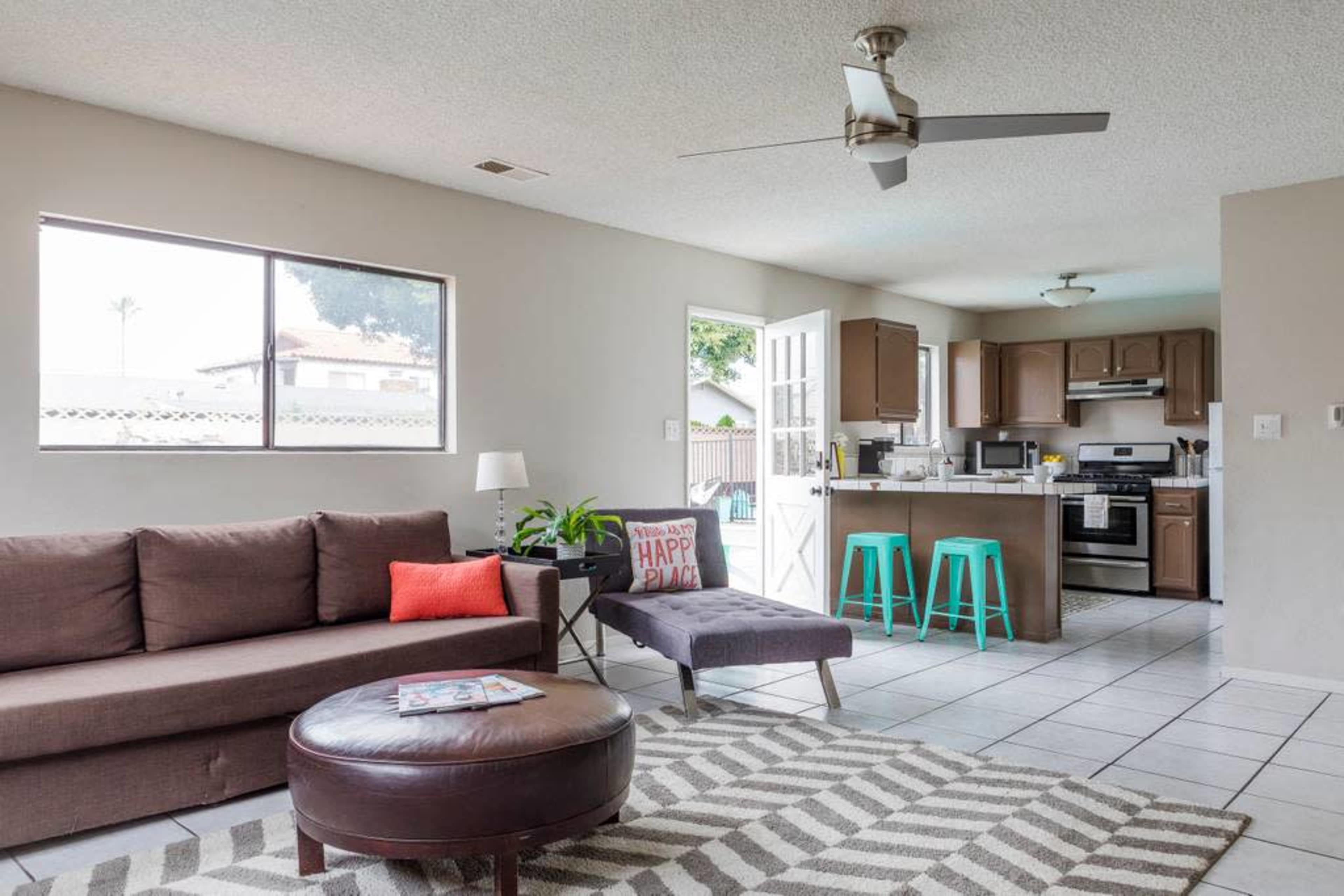 A modern living room with a brown sectional sofa, a round coffee table, a gray chaise lounge, and a kitchen area in the background.