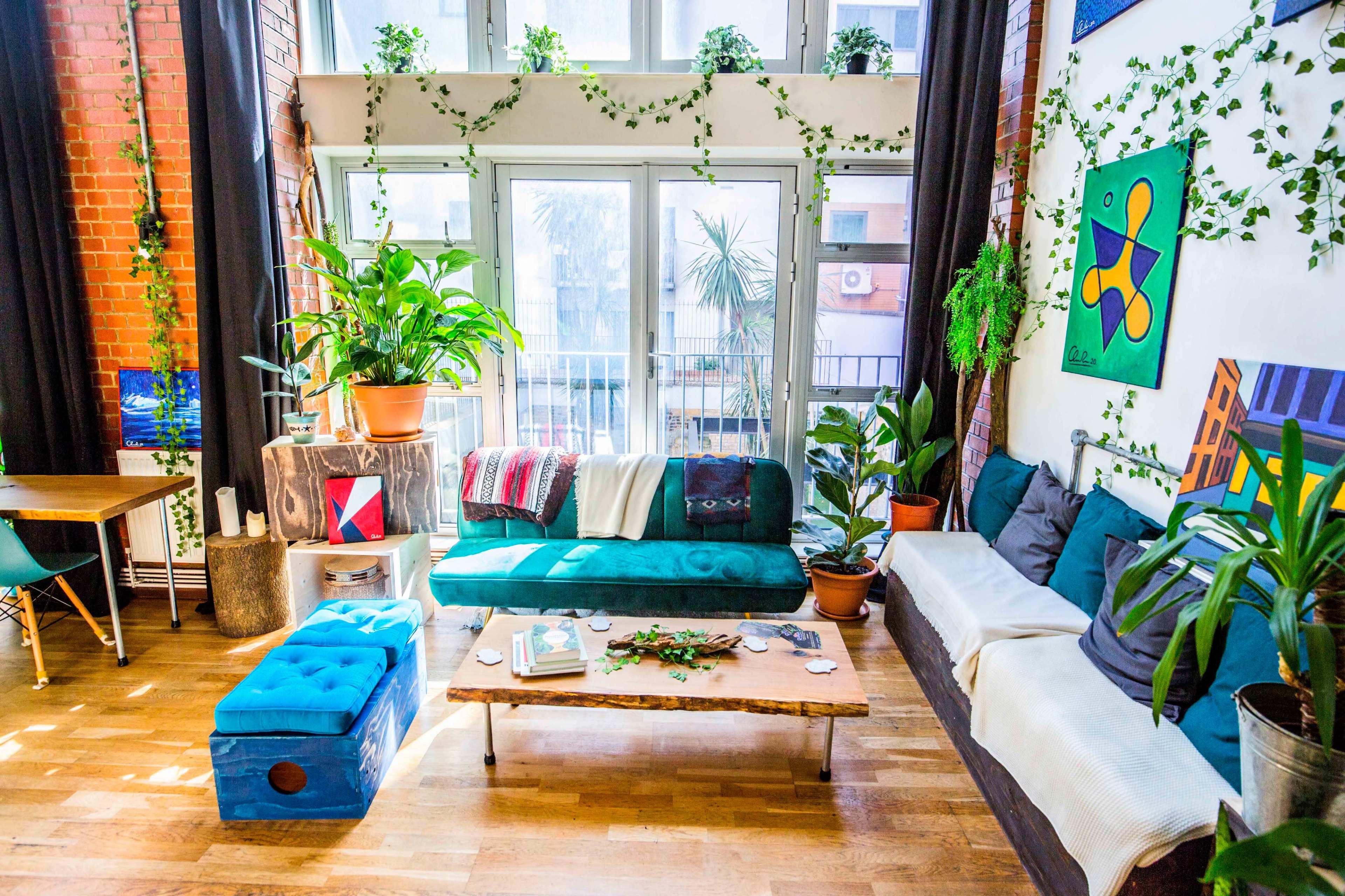 A bright living room filled with plants, featuring a teal couch, wooden coffee table, and large windows allowing natural light.