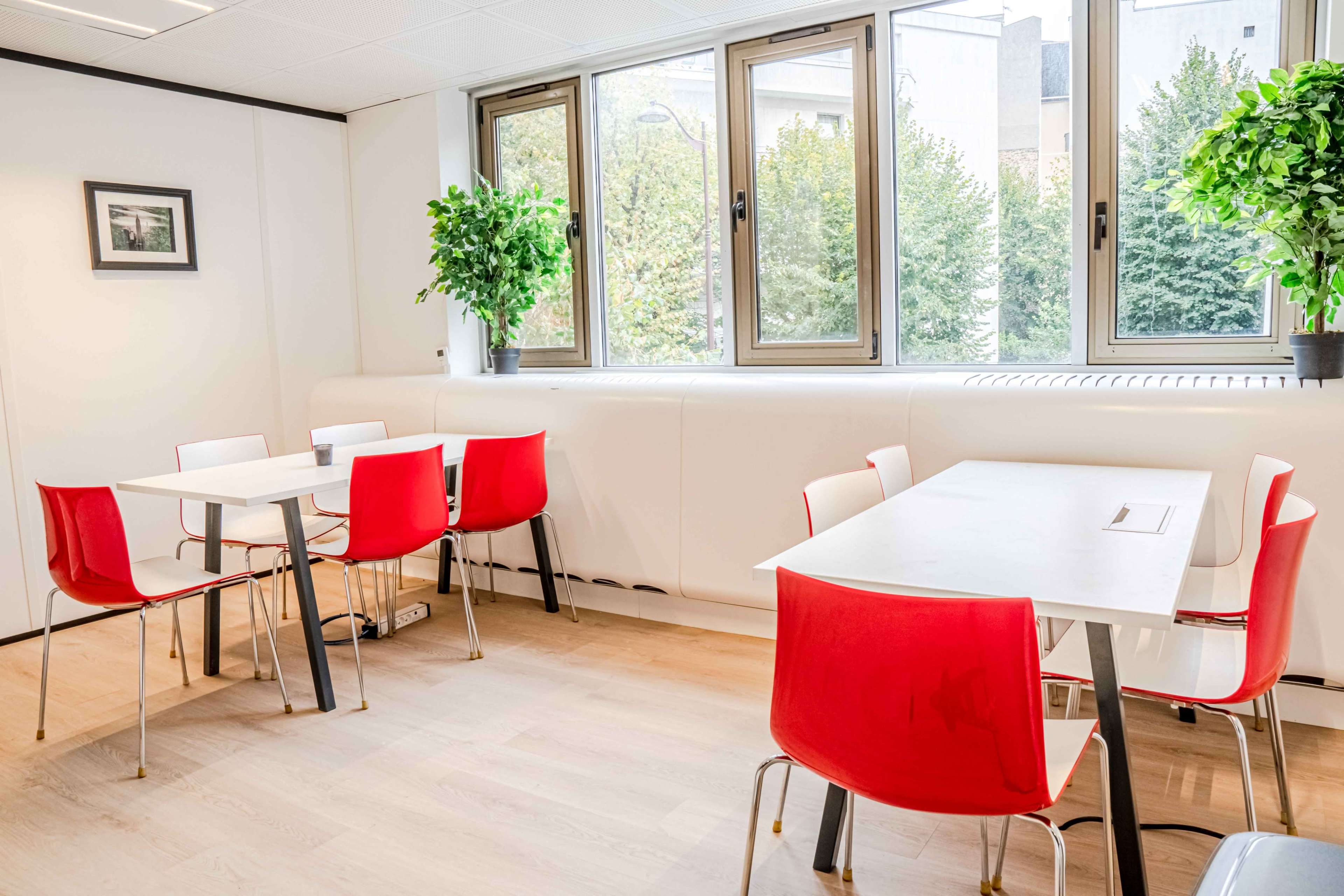 The image shows a bright, modern dining area with white walls, light wooden flooring, red chairs, and tables near large windows.
