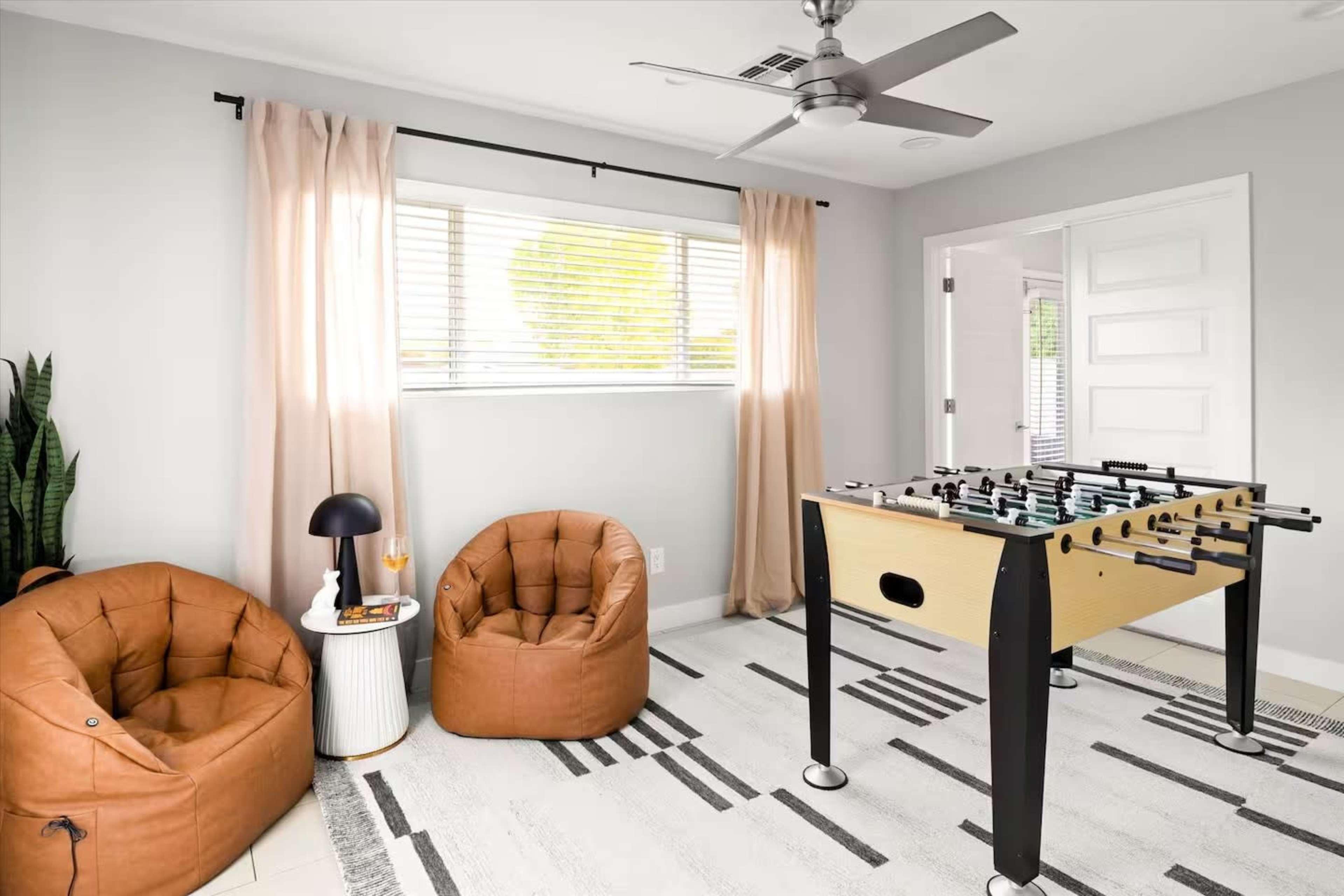 The image shows a bright room with two brown bean bag chairs, a small side table, a foosball table, and a window with light drapes.