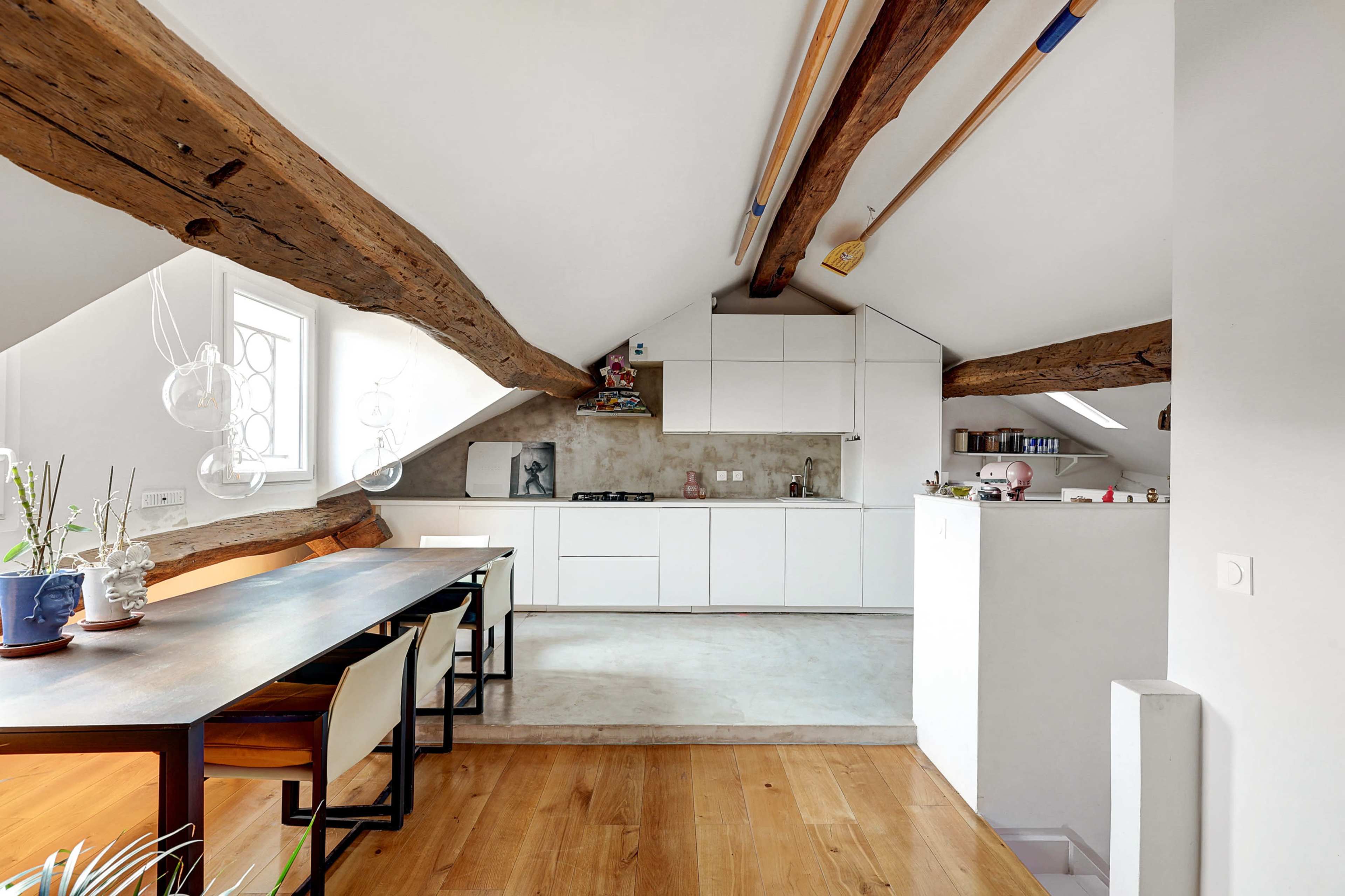 A modern kitchen and dining area with exposed wooden beams, a long dining table, and minimalist cabinetry.