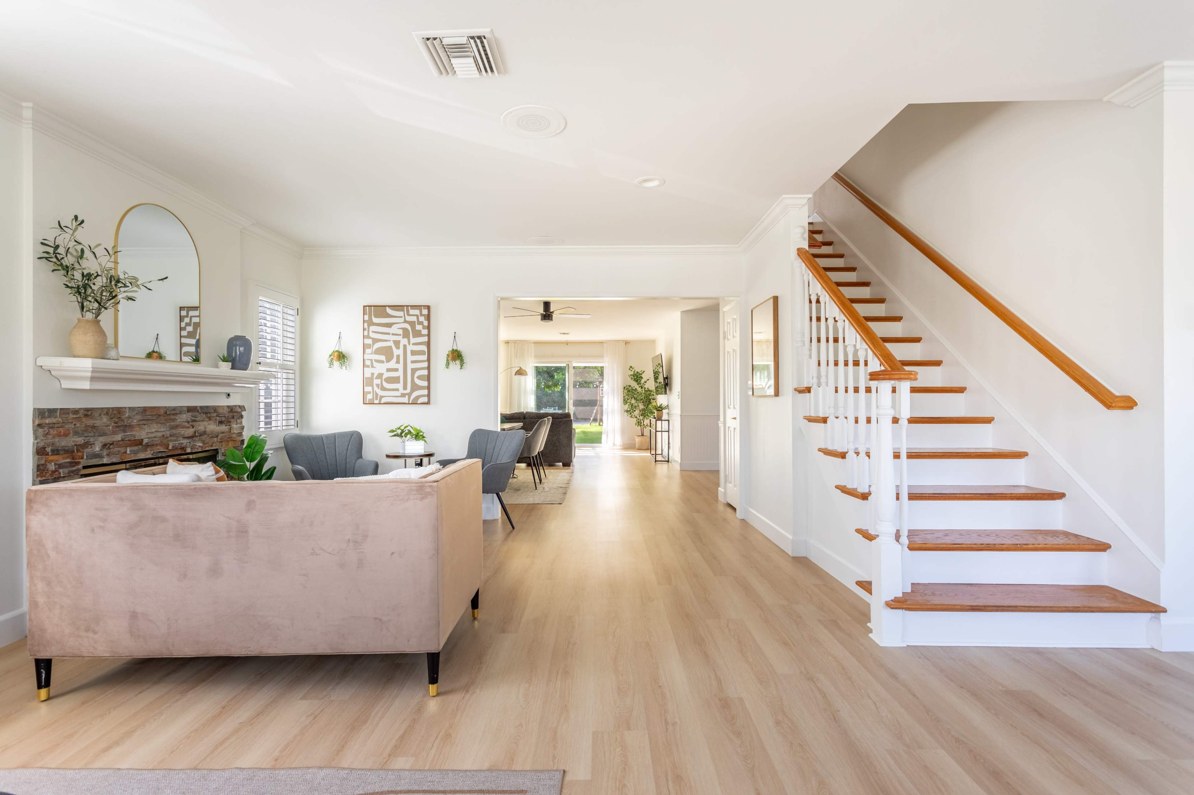 The image shows a spacious living area with a beige couch, a staircase on the right, and a view of an open hallway leading to another room.