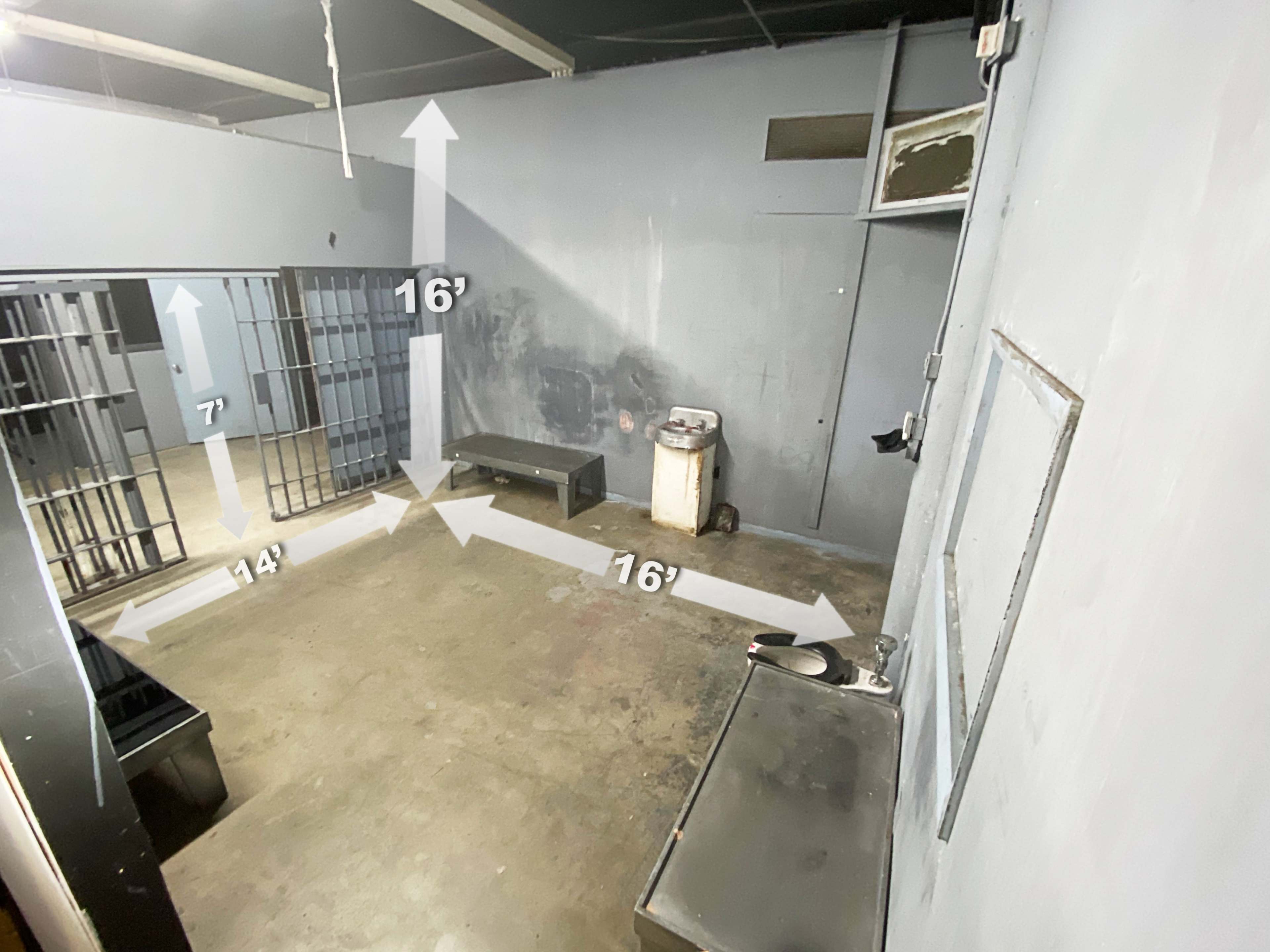 The image shows a sparsely furnished room with gray walls, including two jail cells on the left, a bench in the center, and a sink against the wall, marked with measurements indicating the dimensions of the space.