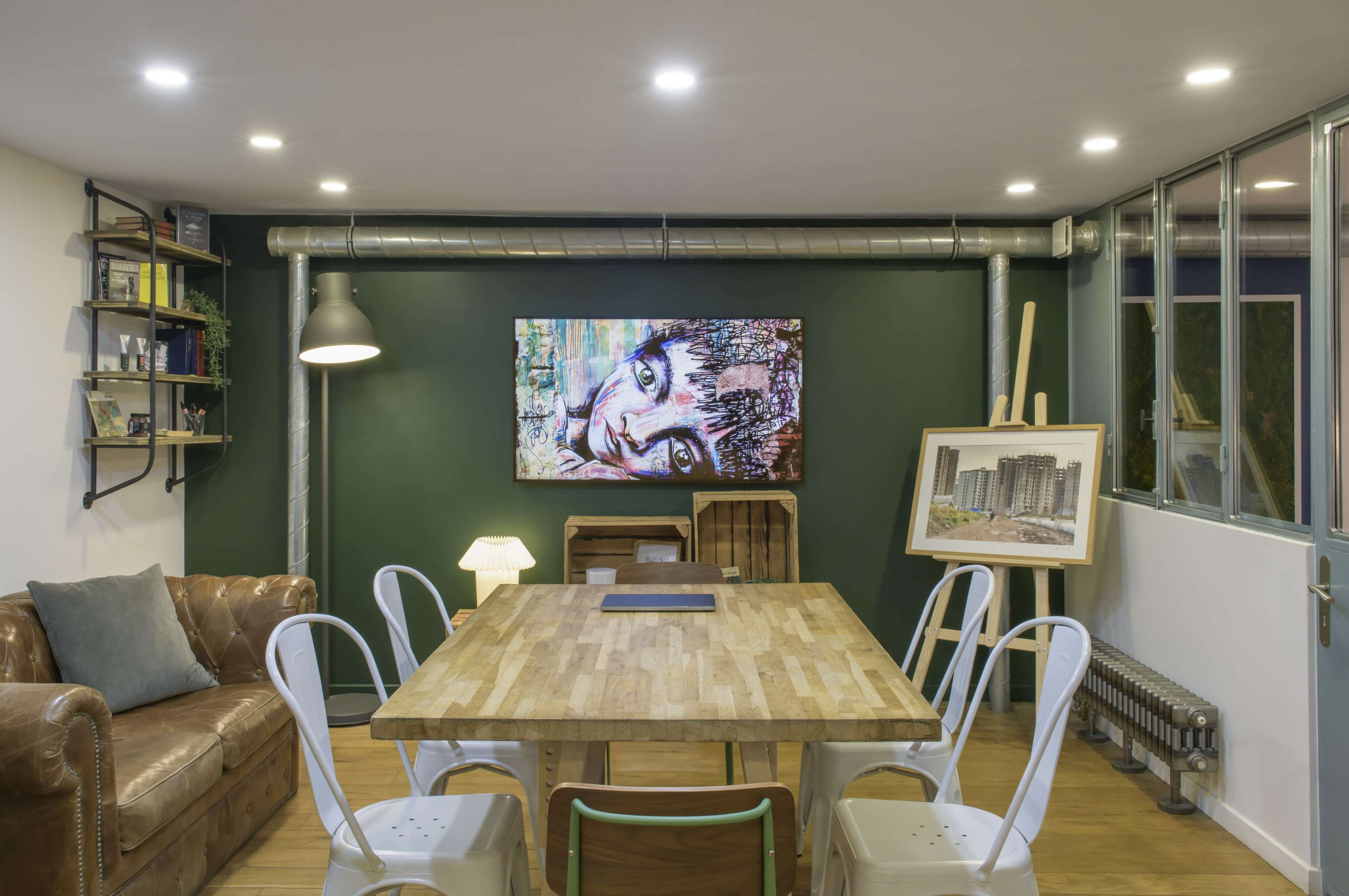 The image shows a modern meeting room with a large wooden table surrounded by white chairs, a brown leather sofa in the corner, and a television displaying artwork on a green wall.