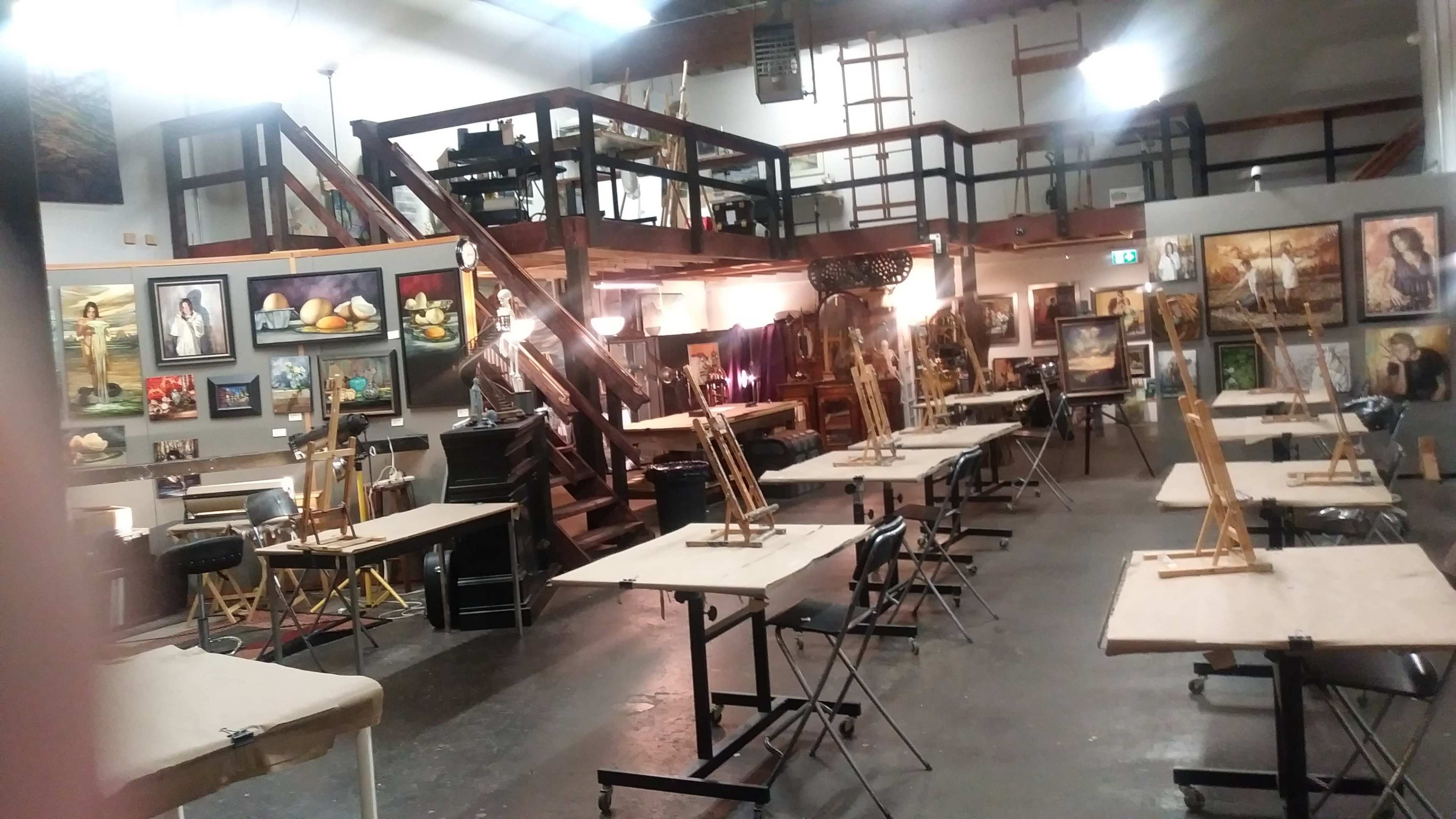 An art studio features several easels arranged around tables, with paintings displayed on the walls and a staircase leading to a loft area.