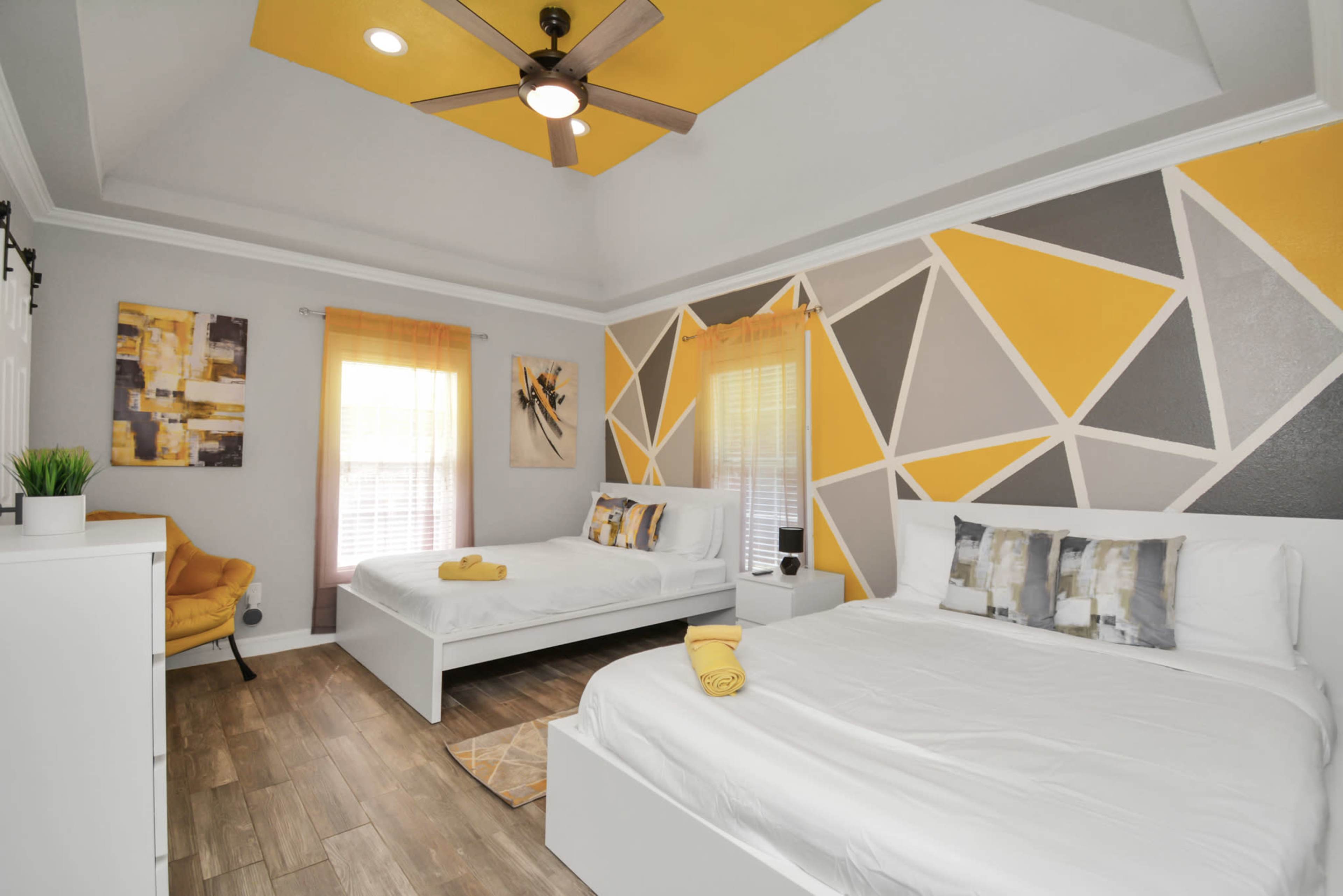 A bedroom featuring two white beds, geometric yellow and gray wall art, and large windows with sheer curtains.