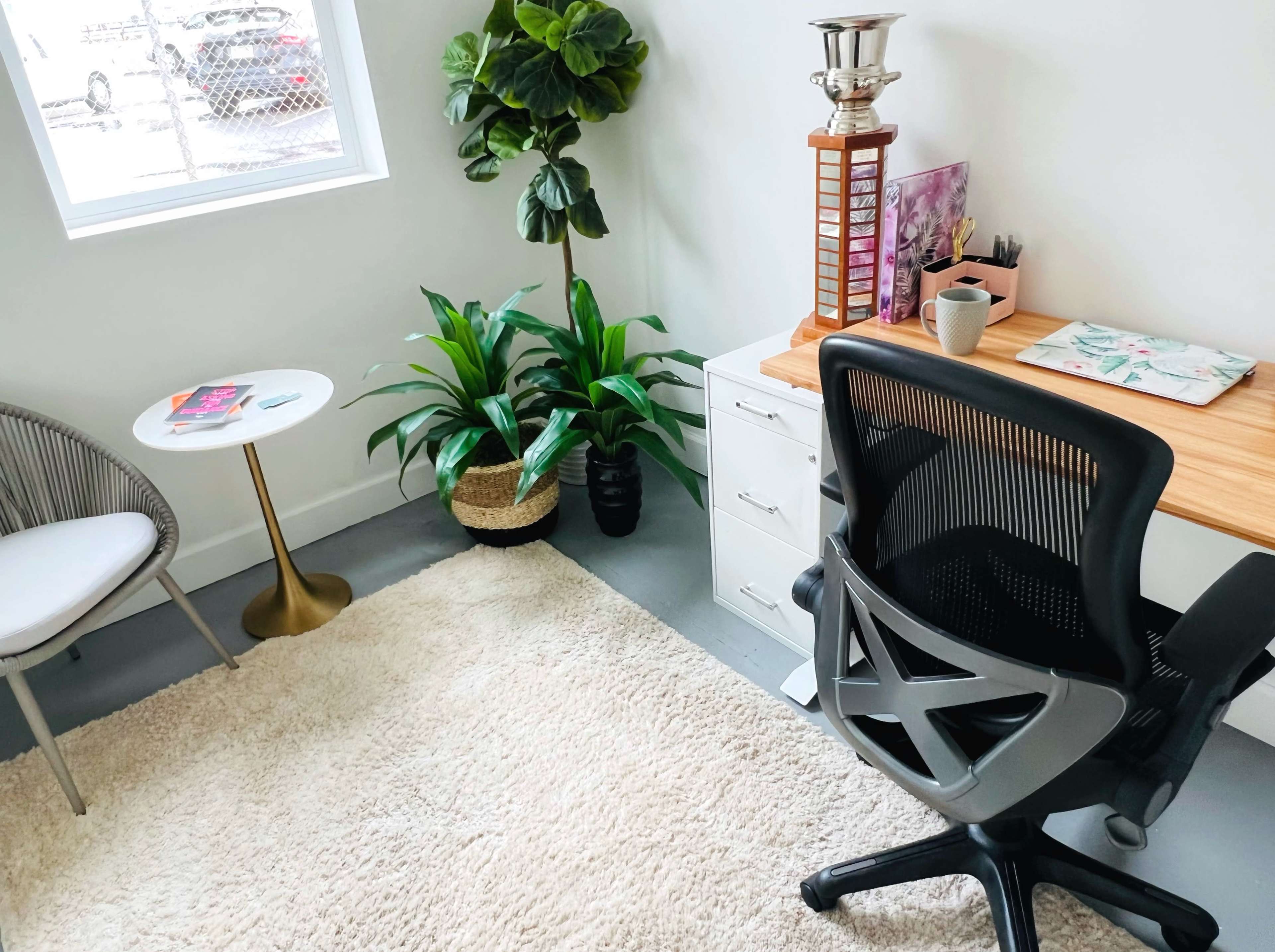 A modern office space features a desk with a wooden top, a black ergonomic chair, a potted plant, a small round side table, and a light-colored rug.