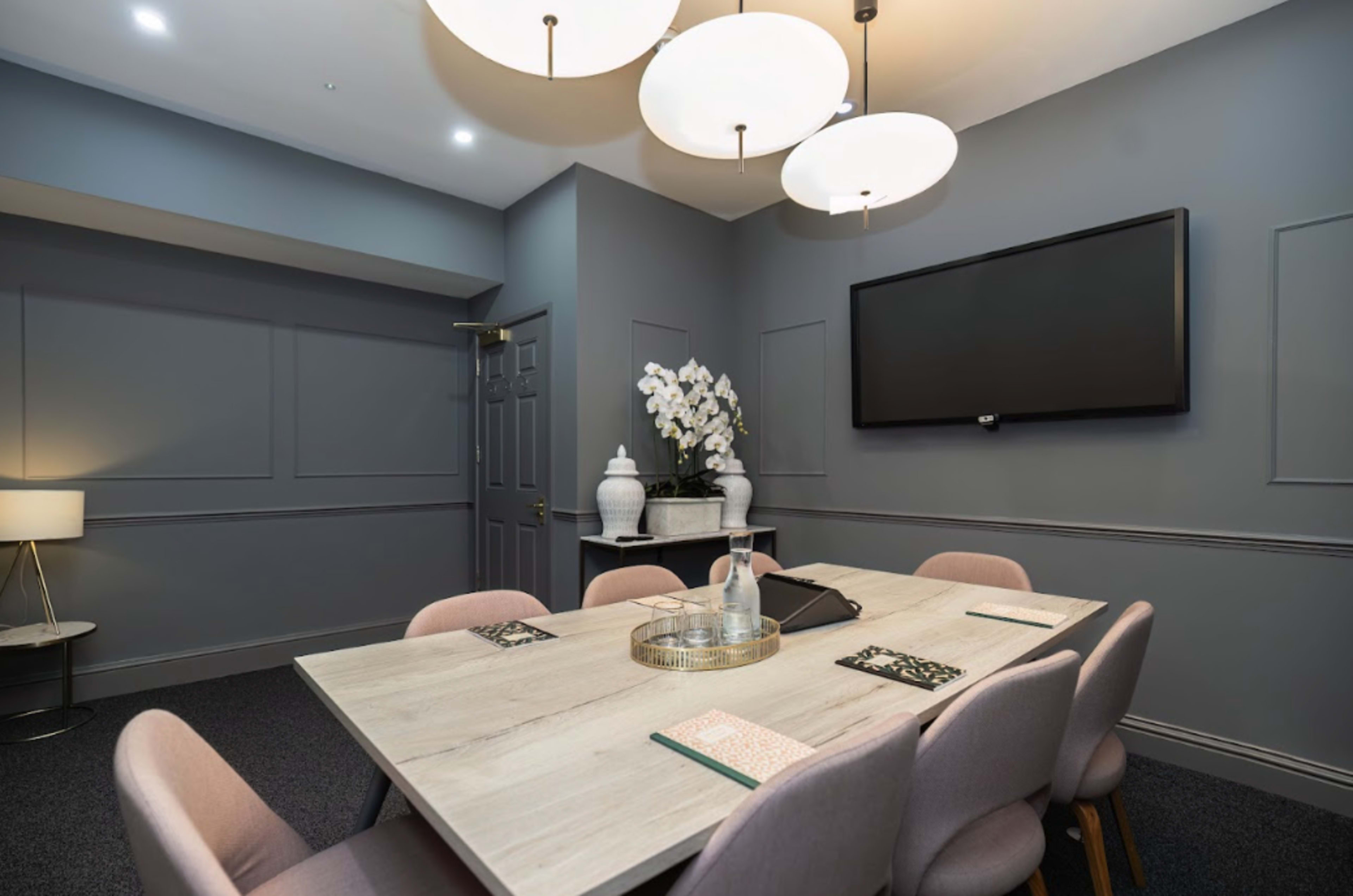 A modern conference room with a large table, six pink chairs, a television mounted on the wall, and decorative plants on a side table.