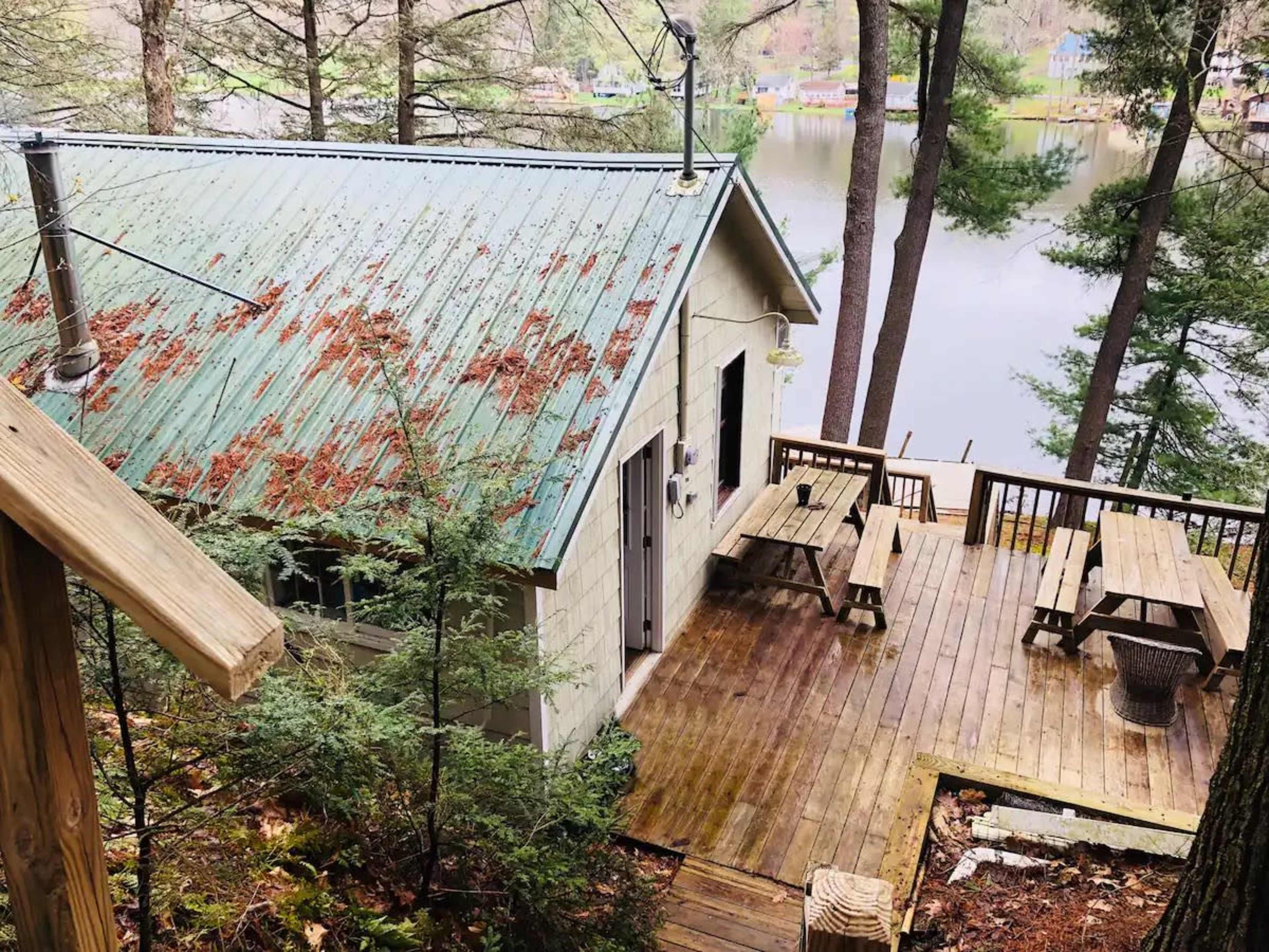 A cabin with a metal roof and wooden deck overlooks a lake, surrounded by trees.