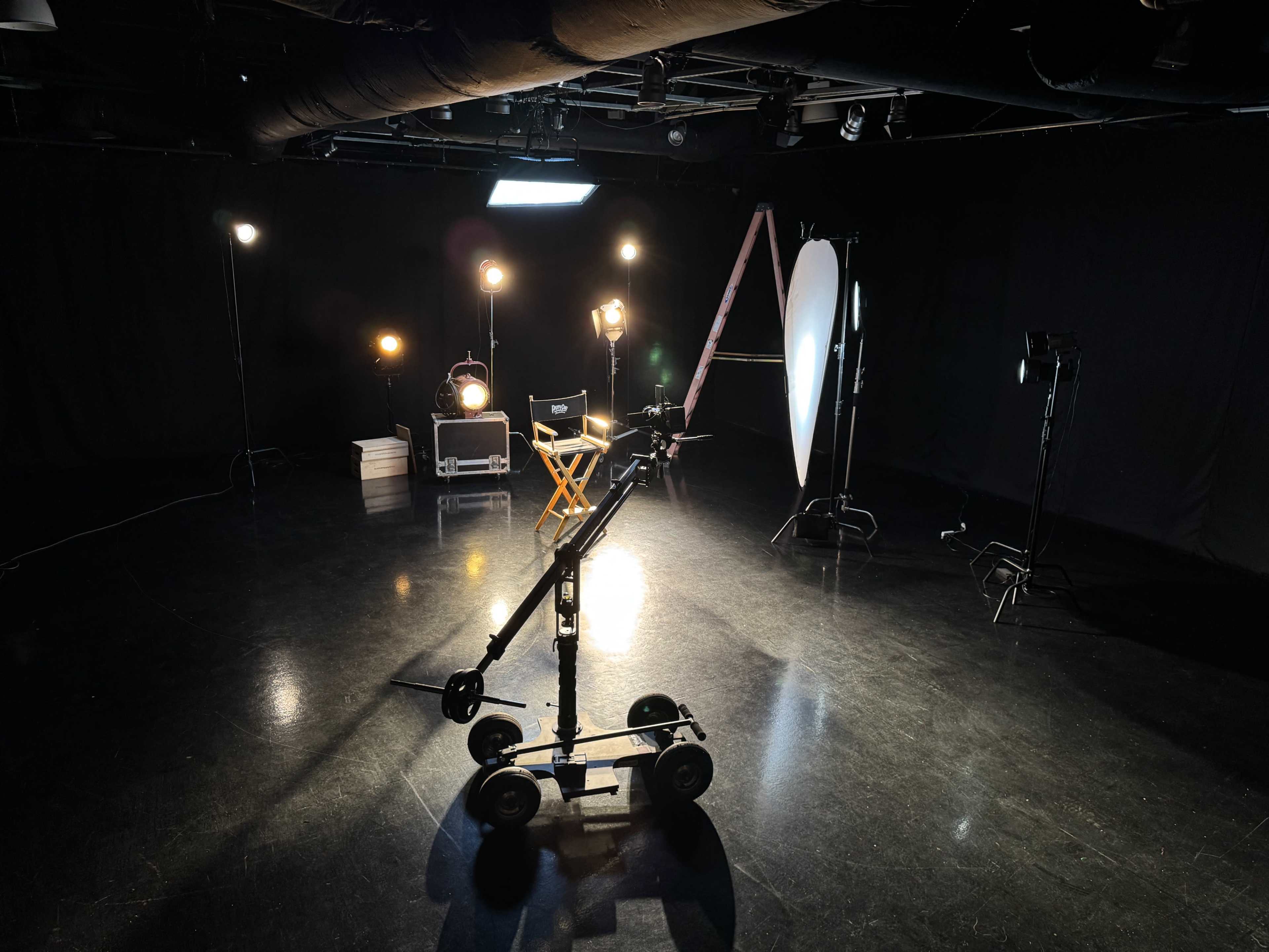 The image shows a dimly lit studio setup with a video camera dolly, lighting equipment, a director's chair, and a reflector.
