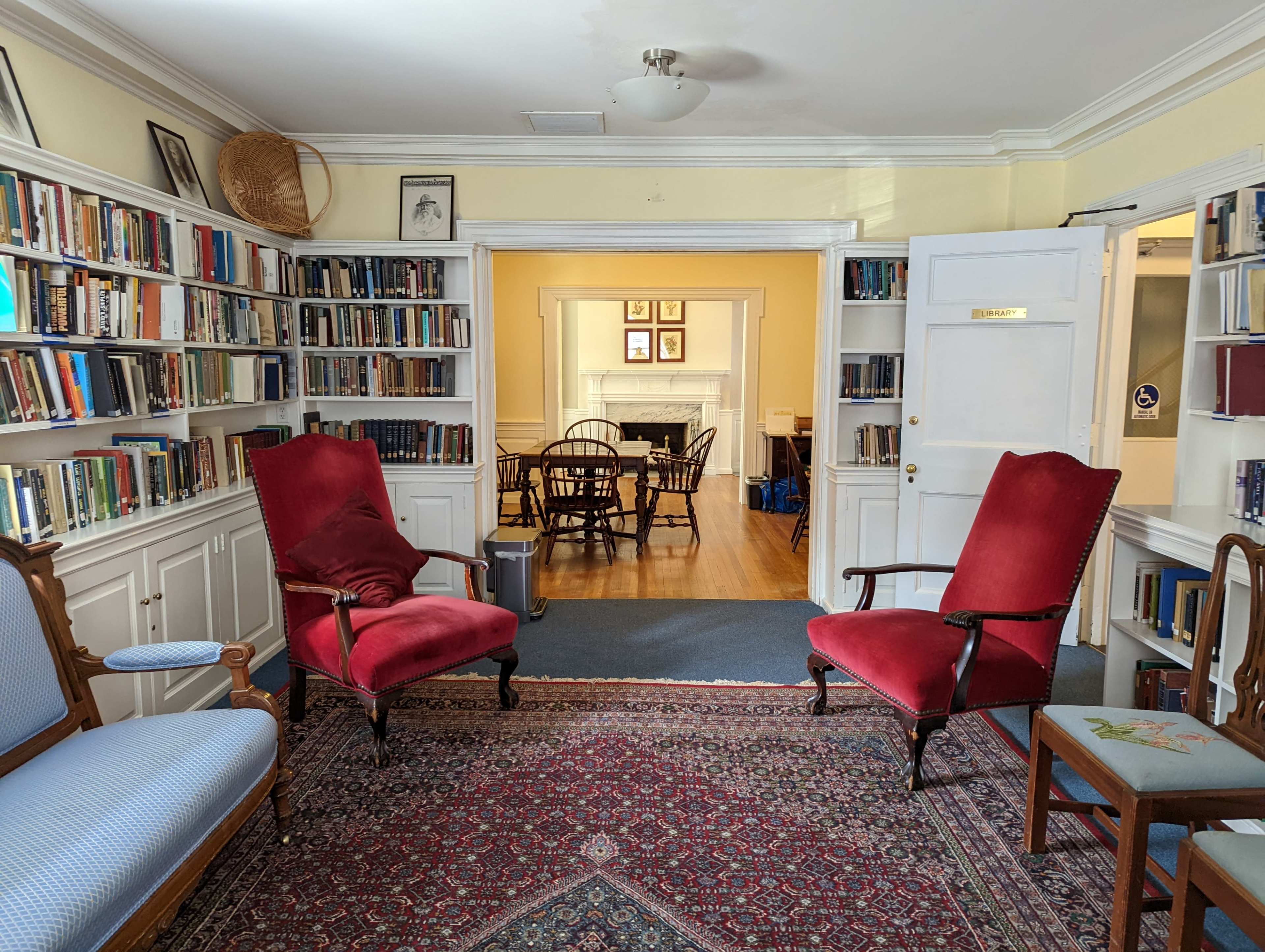 A room furnished with red upholstered chairs, a patterned rug, and bookshelves filled with books lining the walls, leading to a dining area through an open doorway.