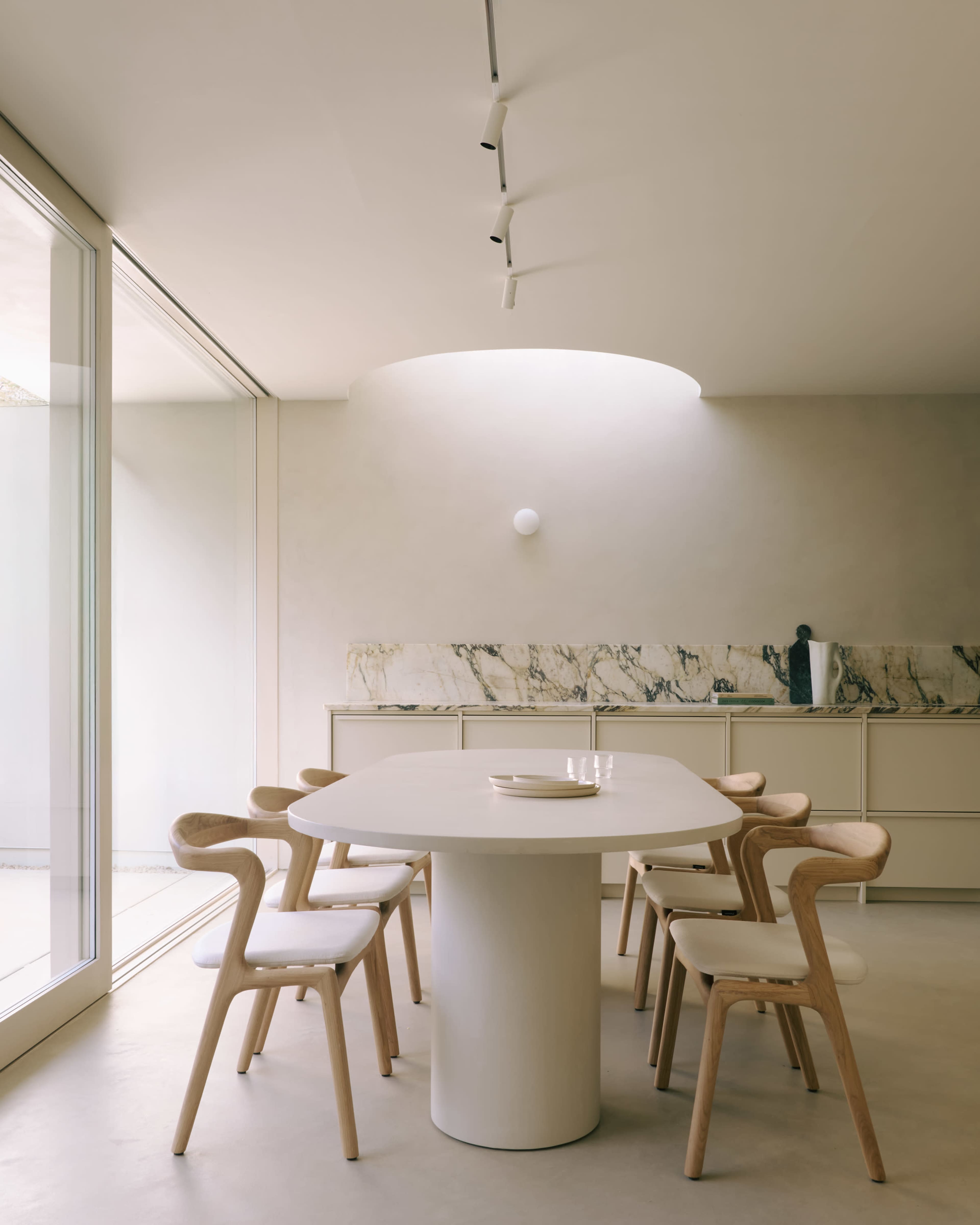 A minimalist dining room features a round table surrounded by wooden chairs, with a marble countertop and large windows letting in natural light.