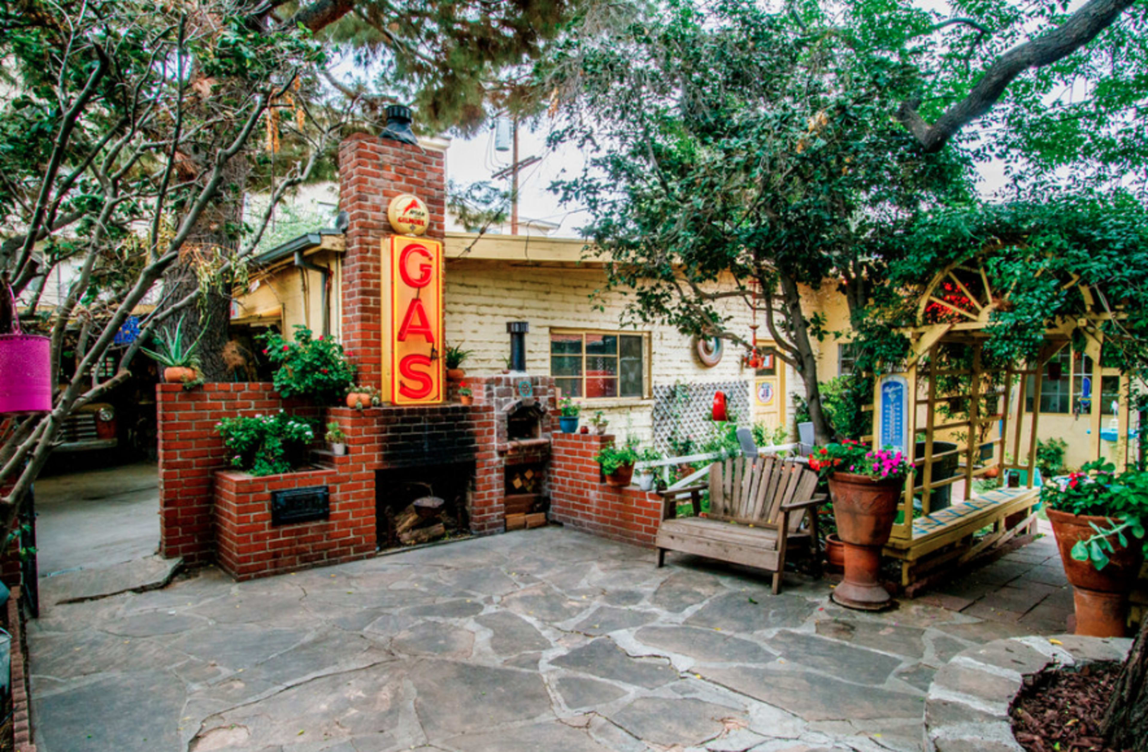 A quaint outdoor area featuring a stone pathway, a brick structure with a vintage gas sign, and a wooden bench surrounded by plants.