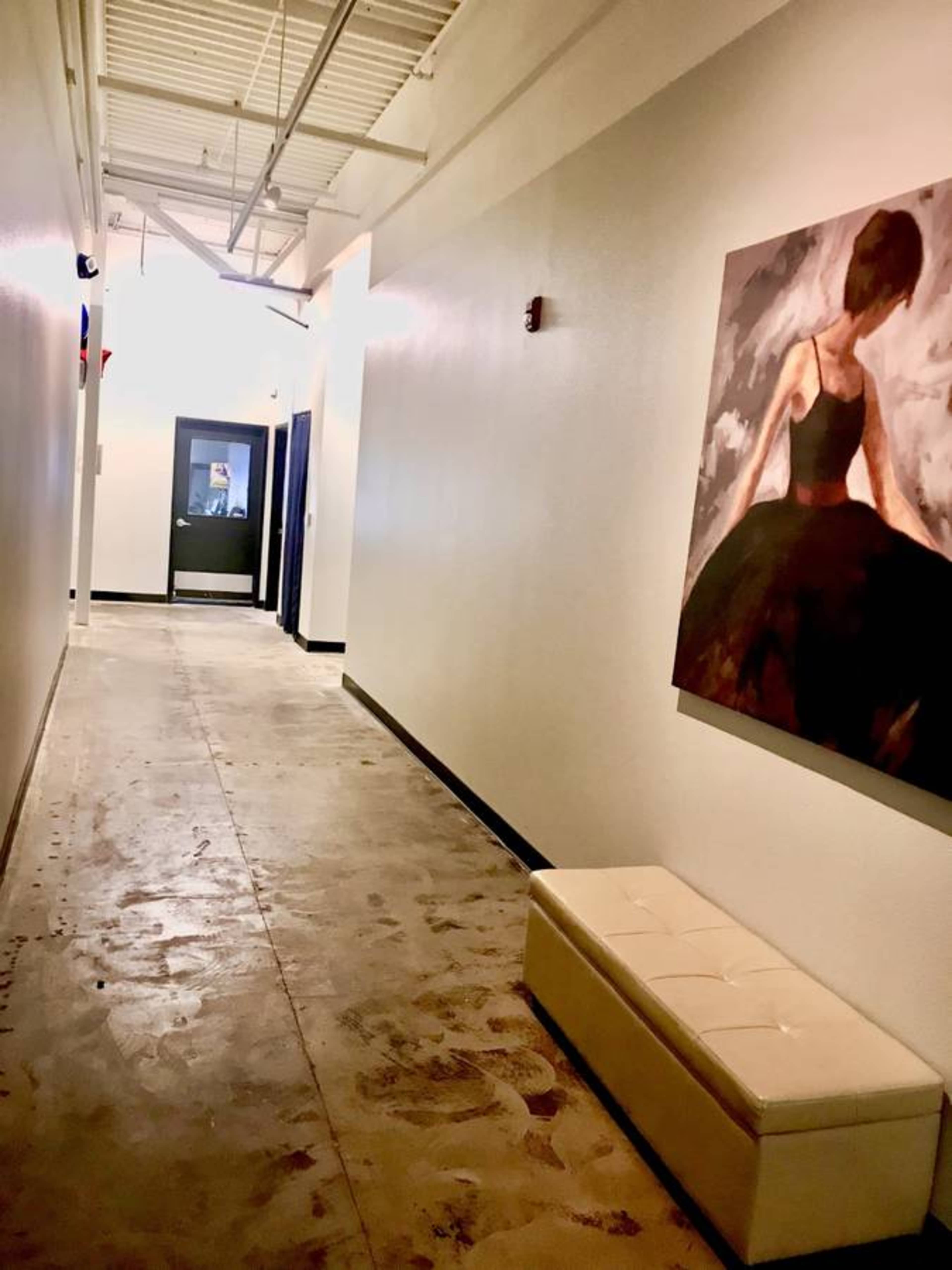 A long, narrow hallway features a white wall with an art piece of a figure in a black dress, and a white bench against the opposite wall.