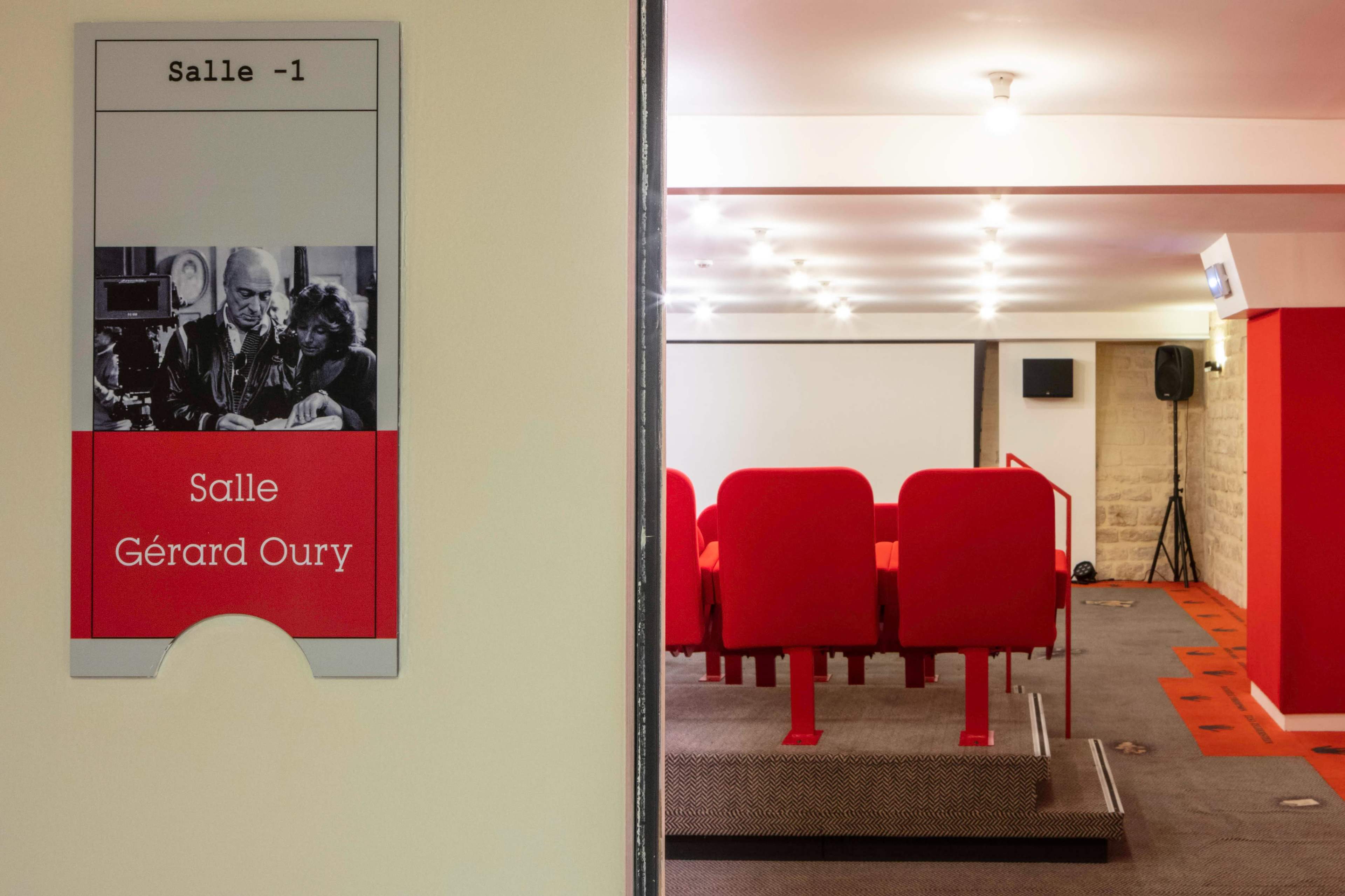 A sign labeled "Salle Gérard Oury" is positioned next to a small theater room featuring red seating and a projector screen.