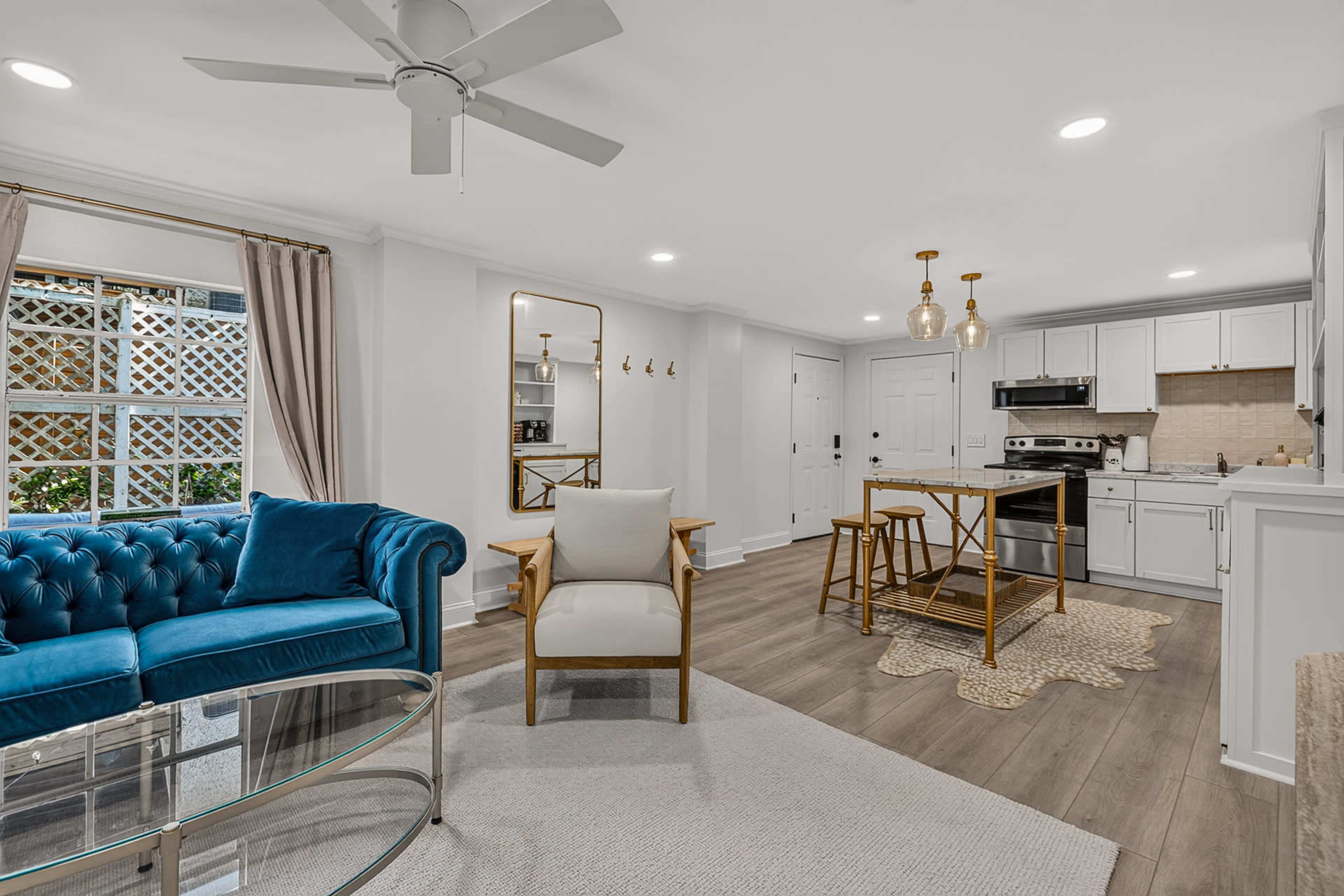 The image shows a modern living room and kitchen area featuring a blue velvet couch, a glass coffee table, a wooden dining table with bar stools, and a well-equipped kitchen.
